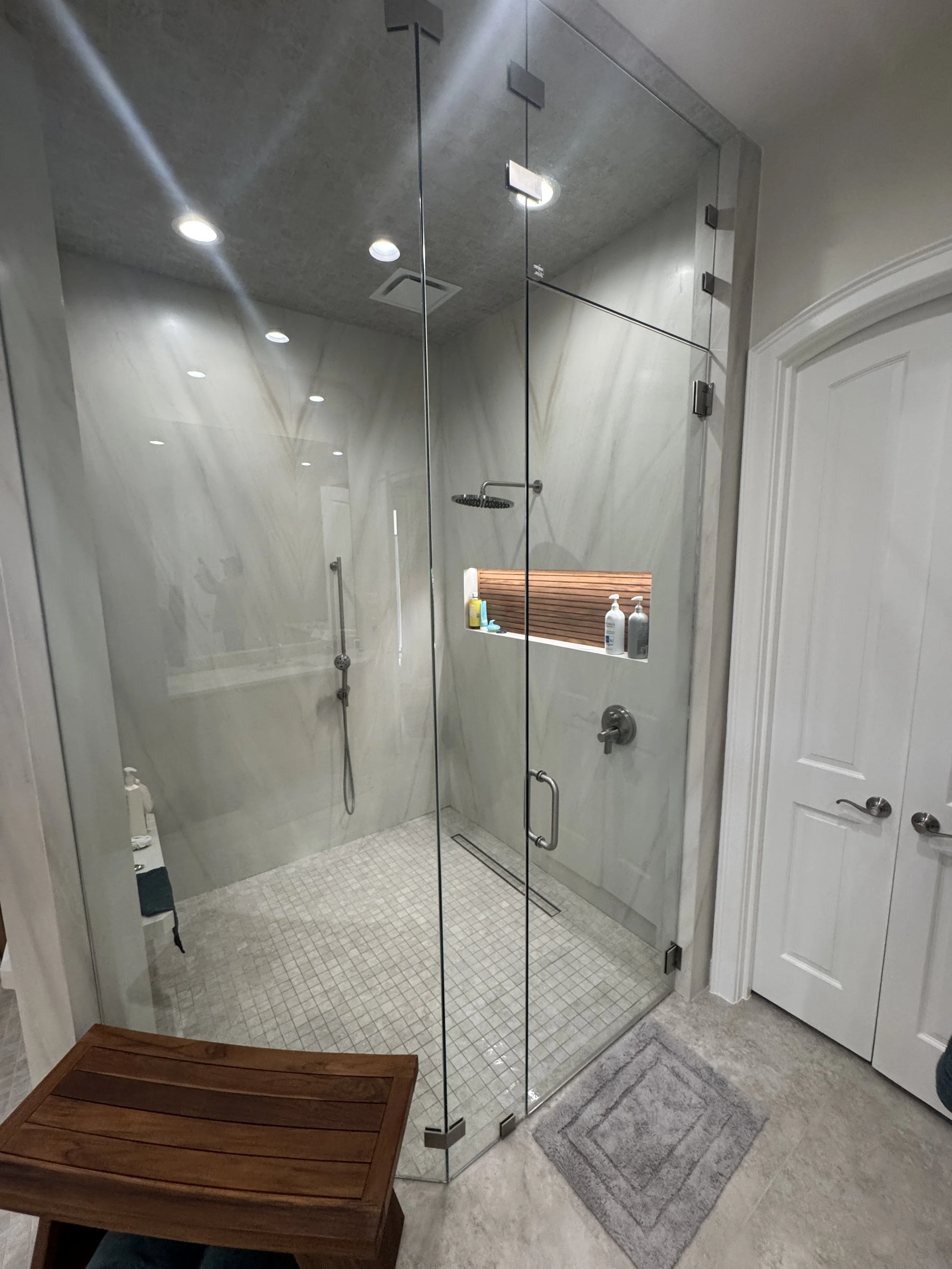 A modern walk-in shower with clear glass doors, a rain showerhead, and a built-in shelf with toiletries, set in a white tiled room with recessed ceiling lights.