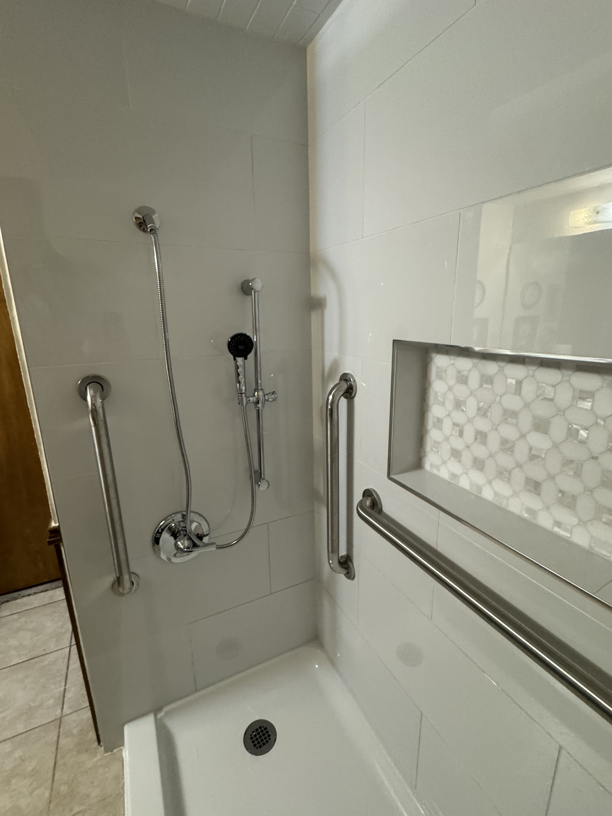 This is a shower stall with beige tile walls, a handheld showerhead, and grab bars for accessibility. It has a built-in tiled seat or shelf, and a drain on the floor.