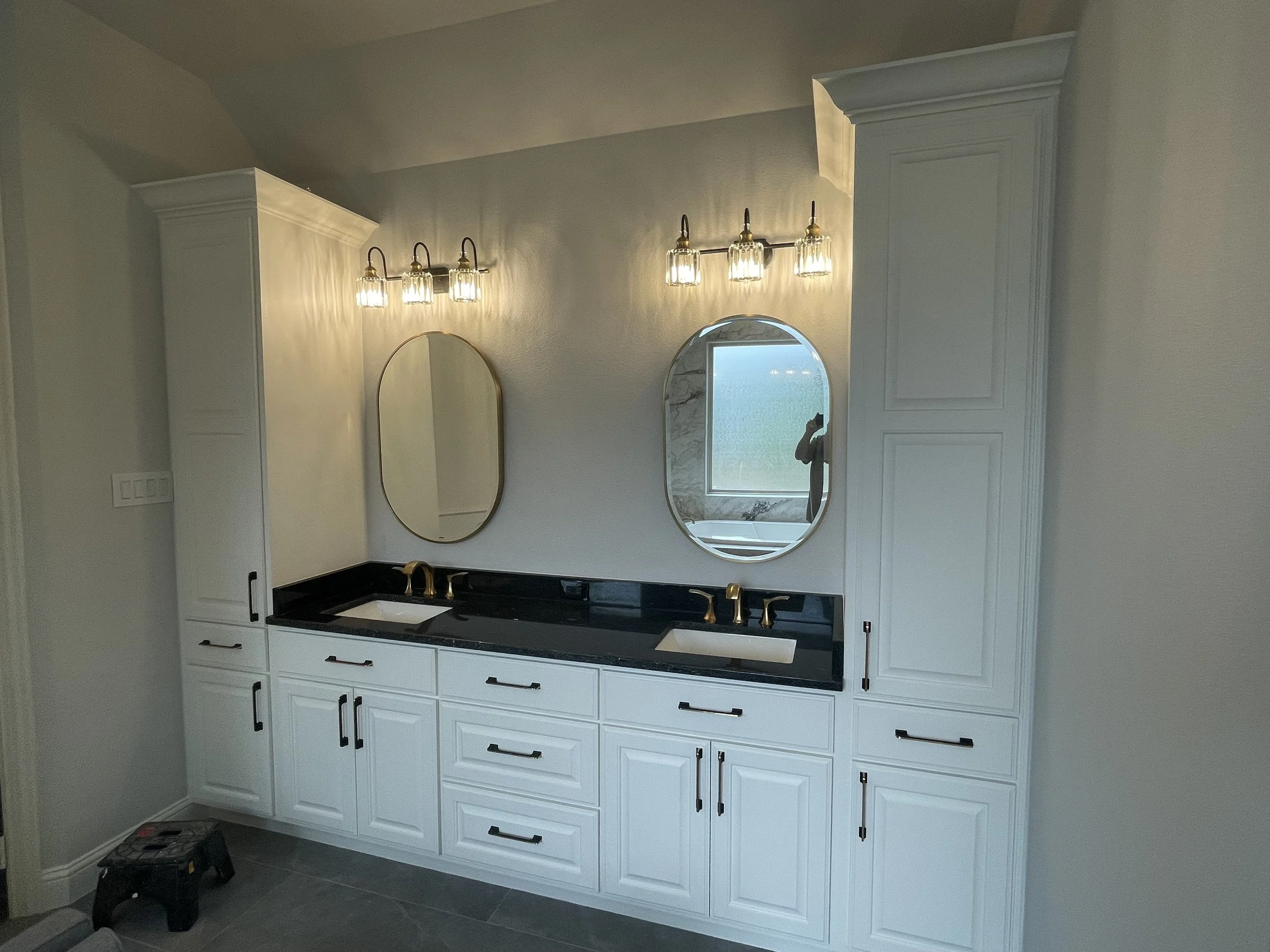 A bathroom vanity with white cabinets, black handles, a black countertop, and a gold faucet. Two oval mirrors are mounted above the sink. Wall-mounted light fixtures with multiple bulbs are on each side of the mirrors. A window with a view of a body of water is in the background.