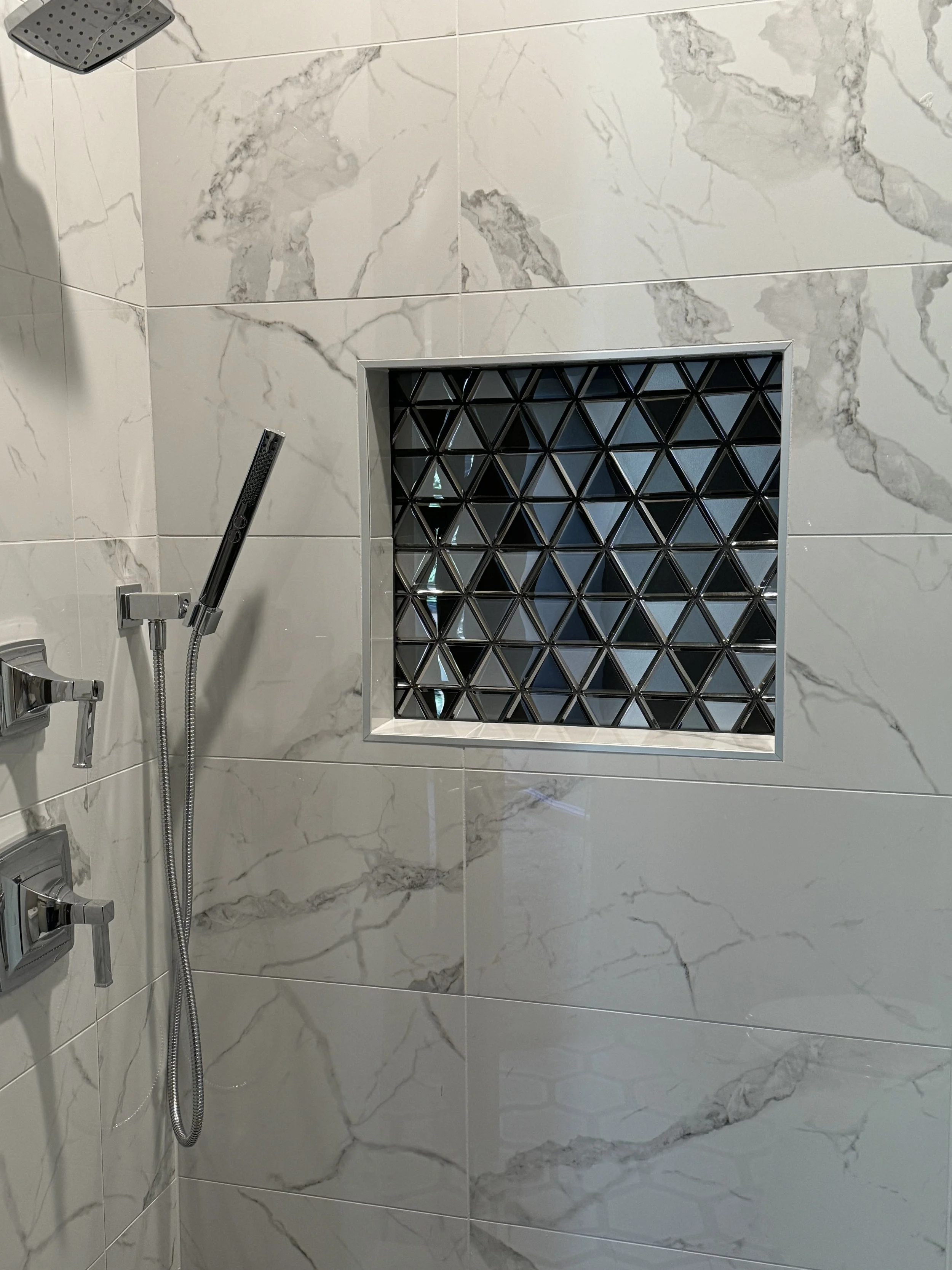 Close-up view of a marble-tiled shower area featuring a built-in recessed shelf with black geometric tile design, a handheld showerhead, and a wall-mounted fixture.