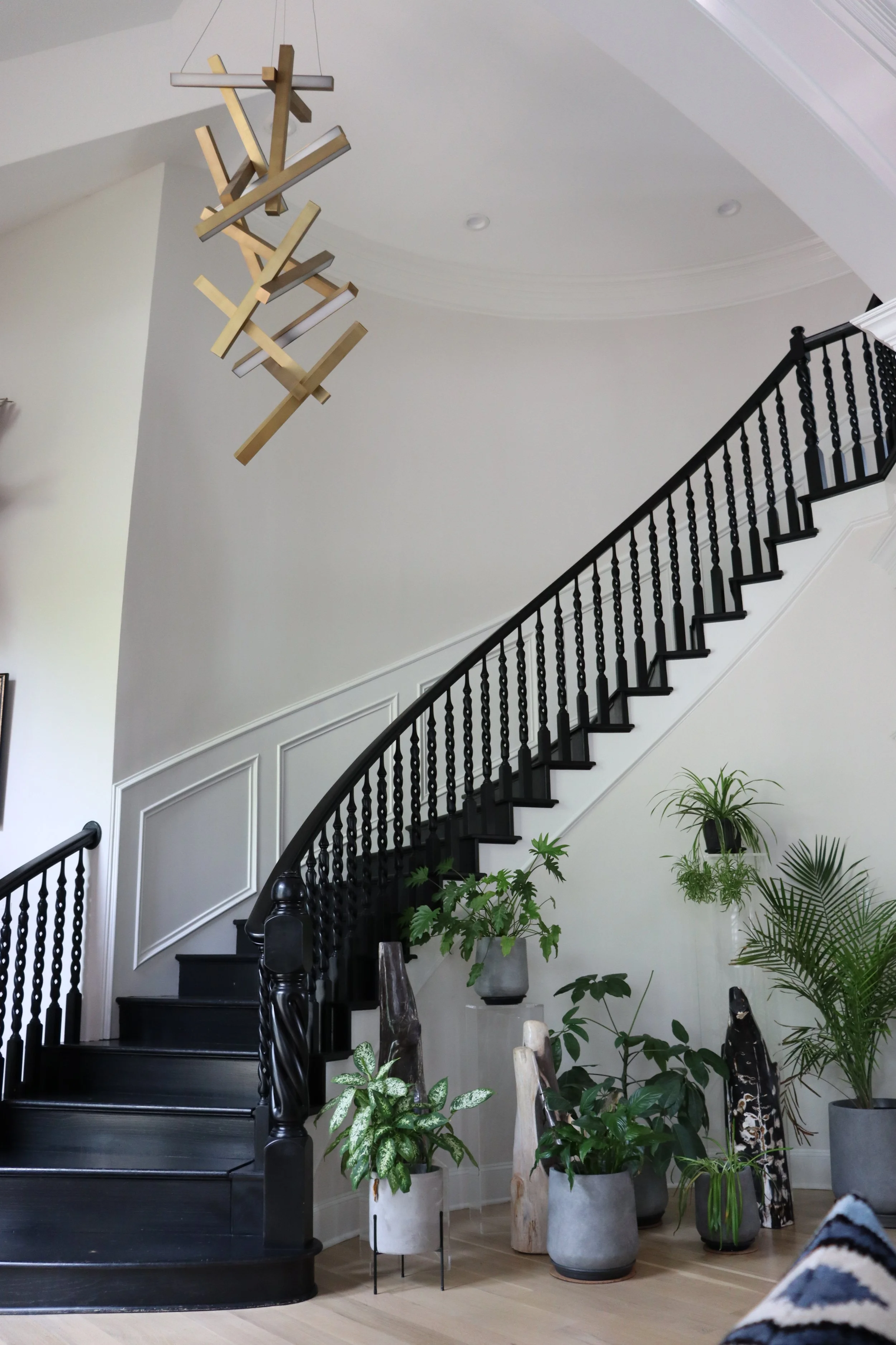 Interior of a home with a black staircase, white walls, and a variety of green plants in decorative pots. A modern chandelier hangs from the ceiling.
