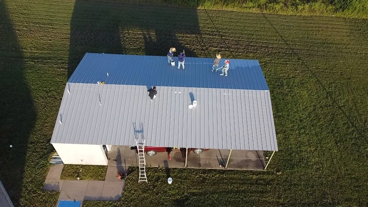 People painting a metal roof, some standing near the edge, others on ladders, with buckets of paint, on a small building surrounded by grass.