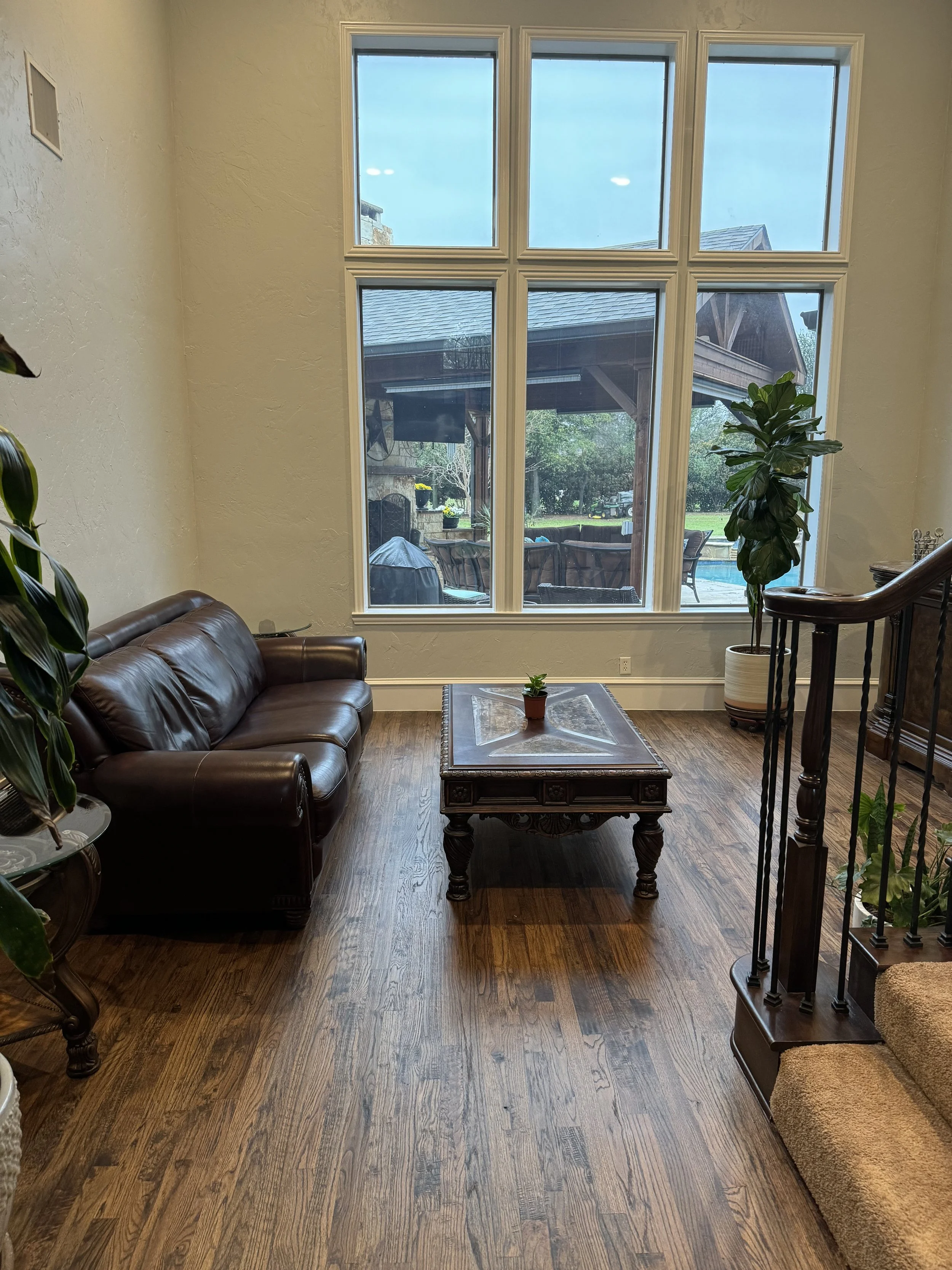 Living room with large window overlooking backyard with outdoor furniture and greenery, brown leather sofa, wooden coffee table, potted plant, staircase railing, and hardwood floor.