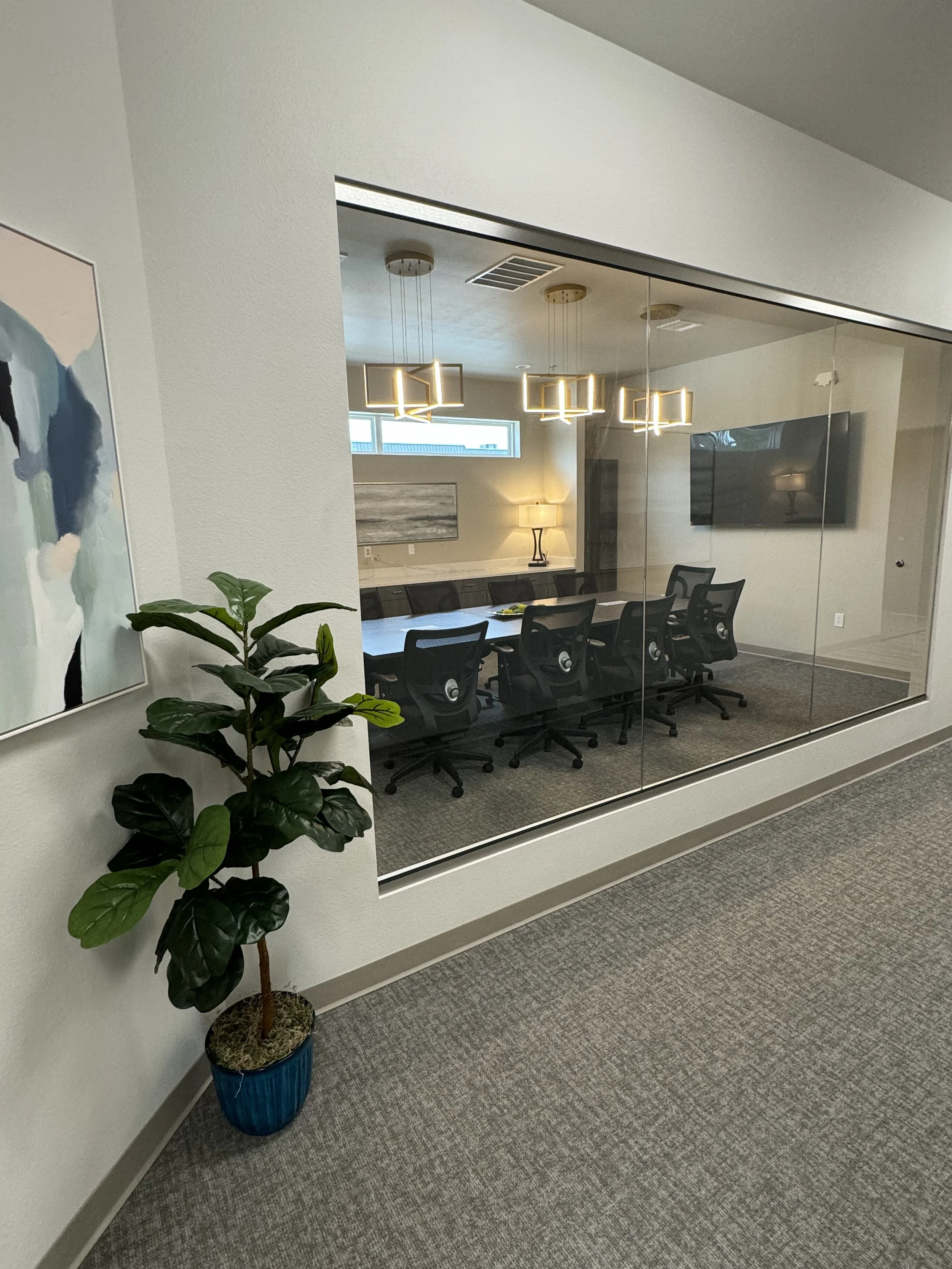 Office conference room with a long table, black chairs, a wall-mounted television, a modern ceiling light fixture, a small window, a nearby wall lamp, a painting, and a potted plant in the foreground.