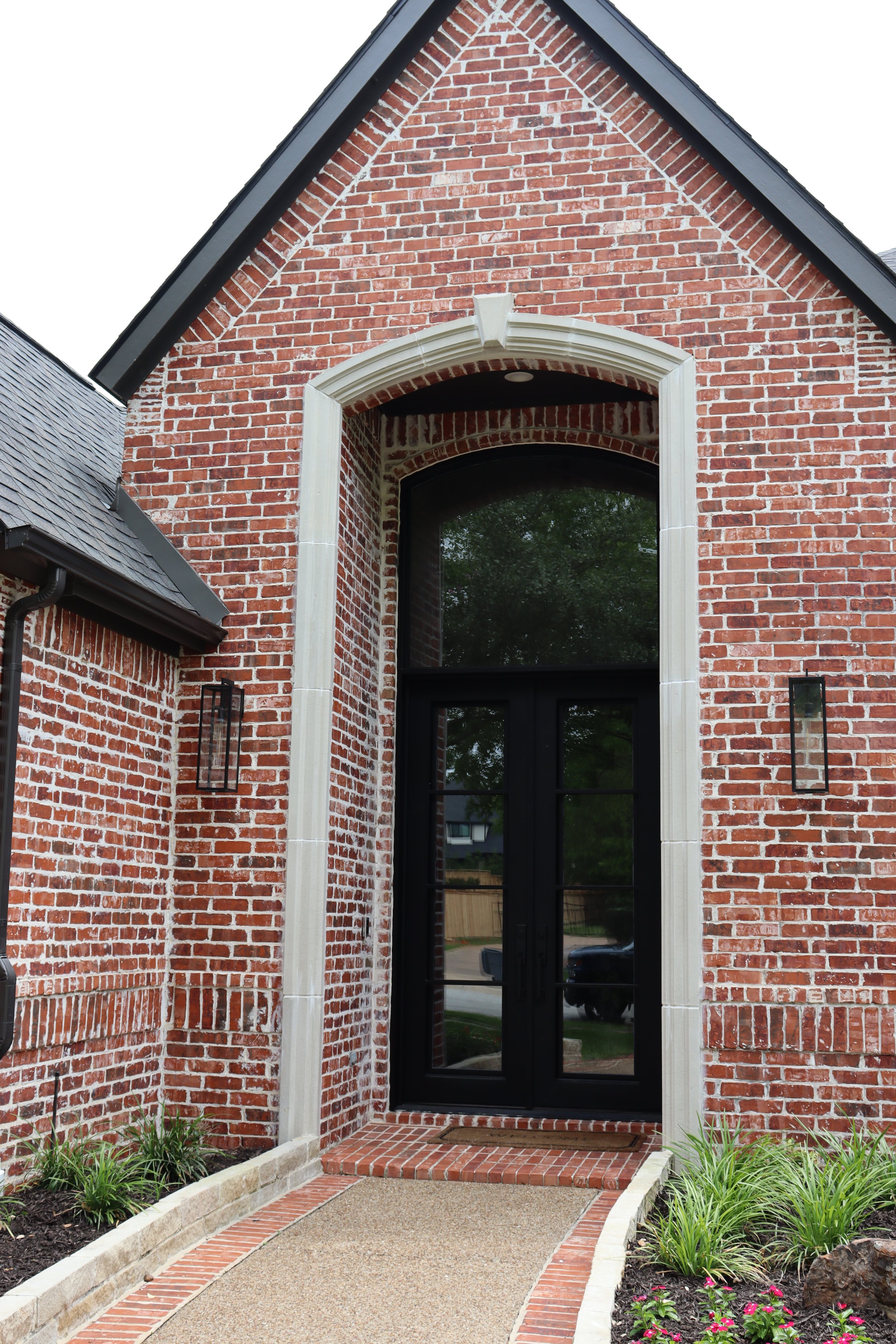 The image shows the front entrance of a brick house with a black door and arched window above it. There is a brick pathway bordered by flower beds with green plants and pink flowers leading to the door, and outside lights are mounted on the wall.