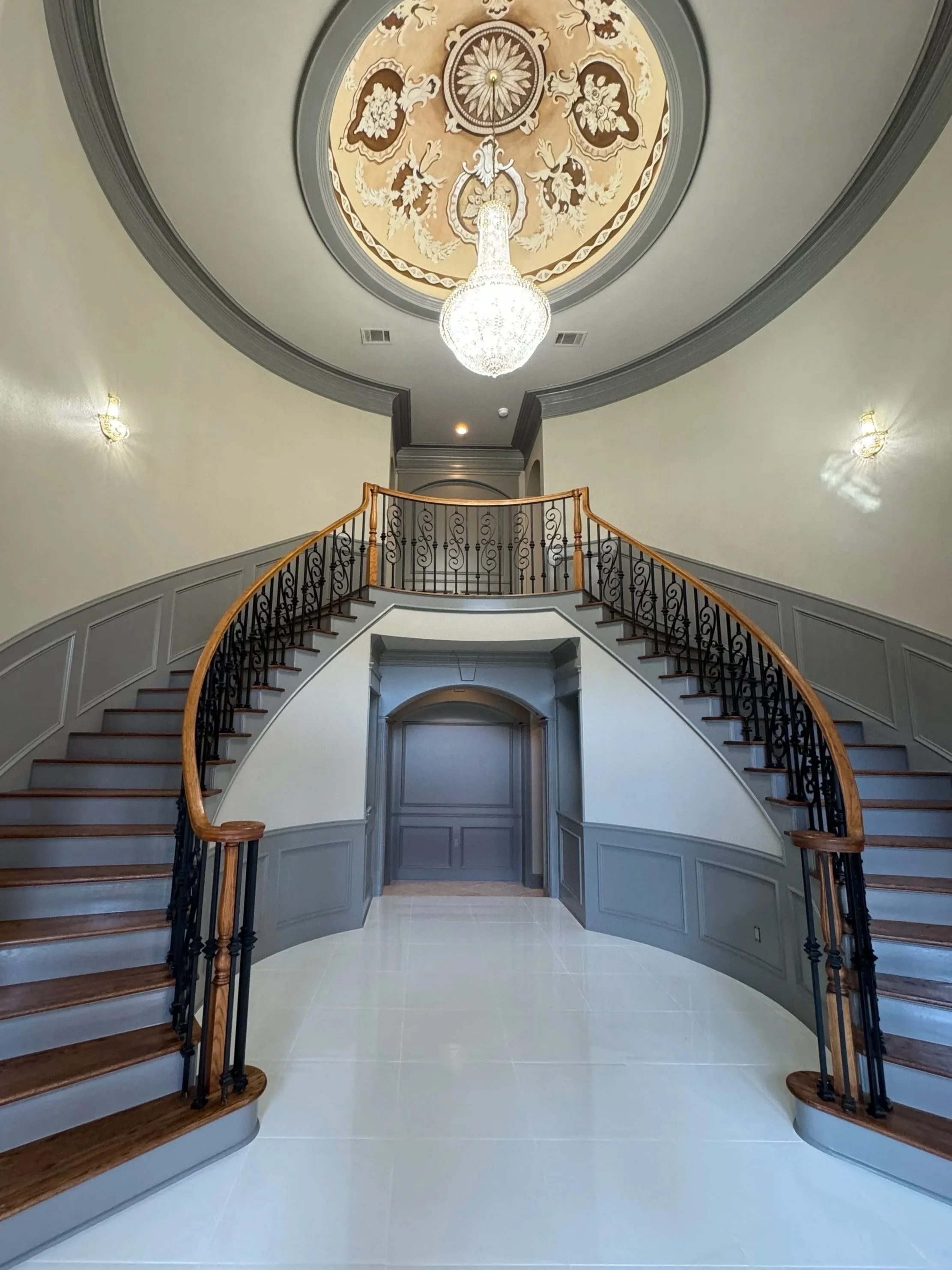Elegant staircase with wooden handrails and black wrought iron balusters, leading to a landing with a decorative ceiling featuring a chandelier and intricate gold and cream patterns, in a spacious hallway with light-colored tiled floor and arched doorways.