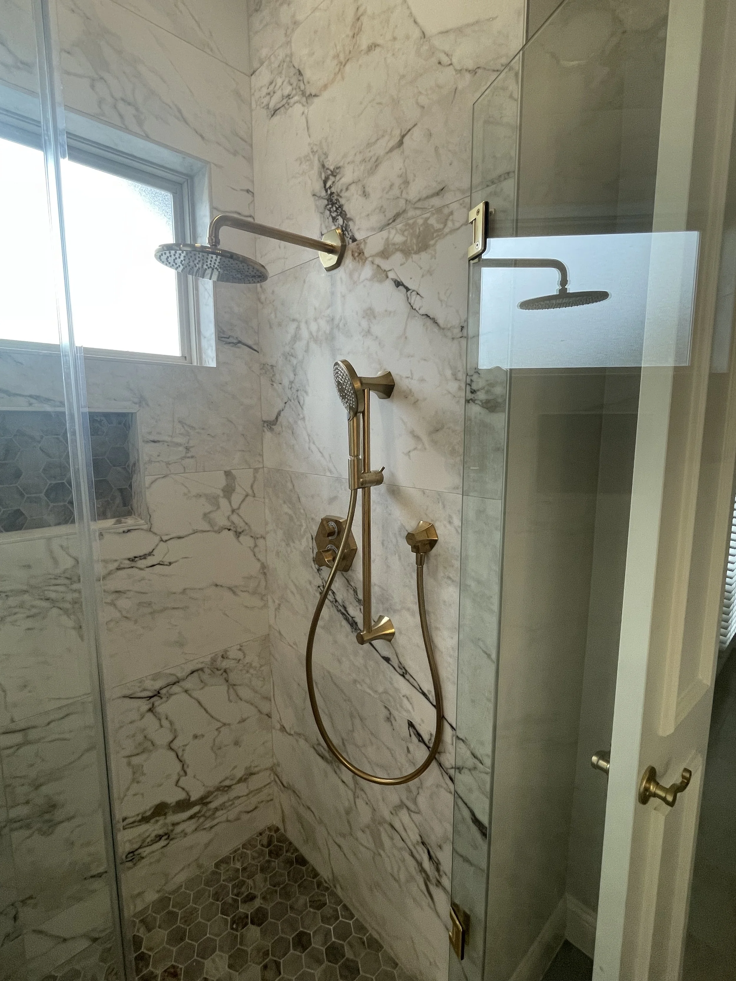 A walk-in shower with marble walls, a window, two brass showerheads, a glass door, and hexagonal tiles on the floor.