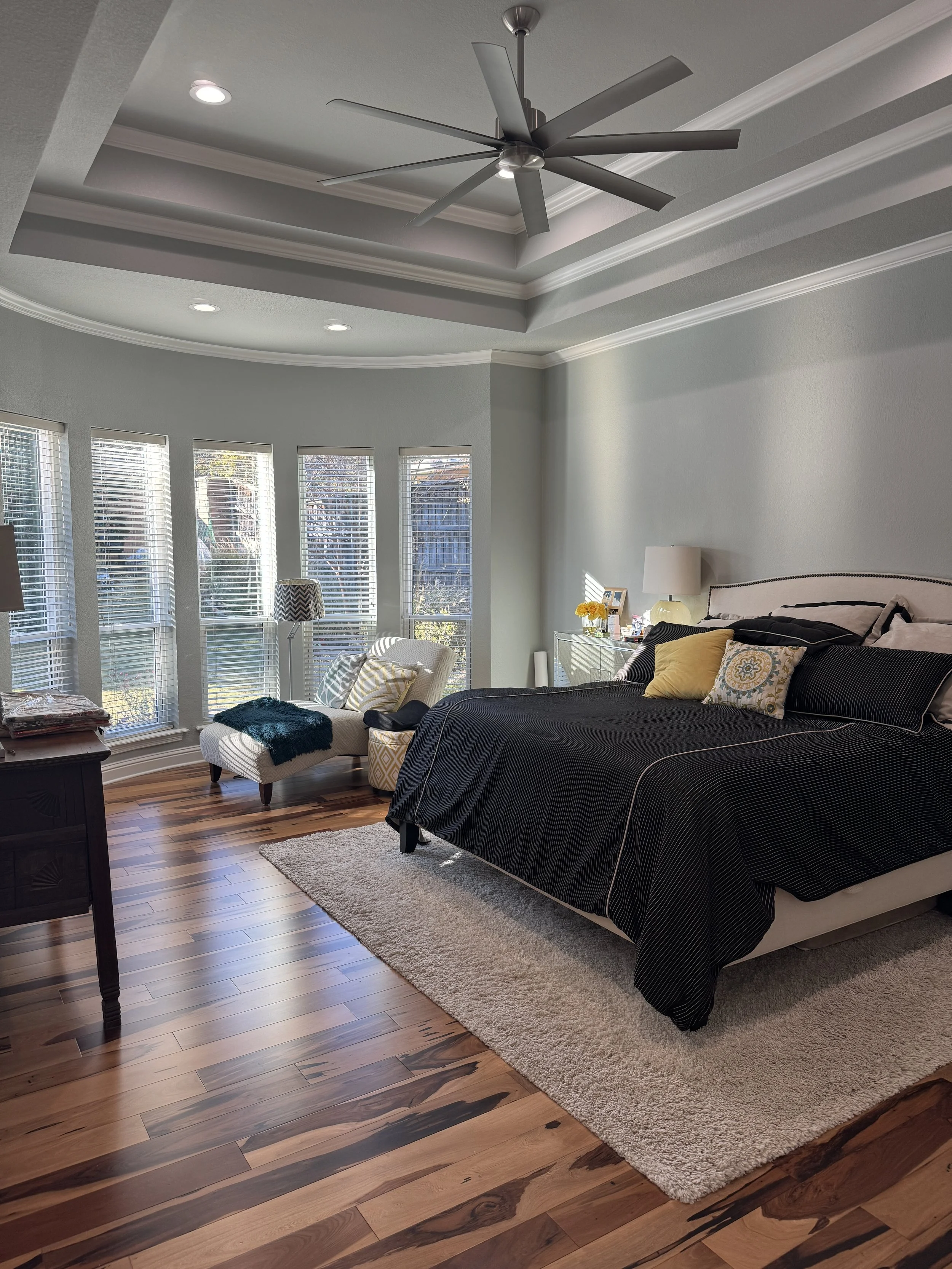 A bedroom with a high ceiling, ceiling fan, multiple windows, a bed with black bedding, yellow and patterned pillows, a nightstand with a lamp, and a plush rug on hardwood floor.