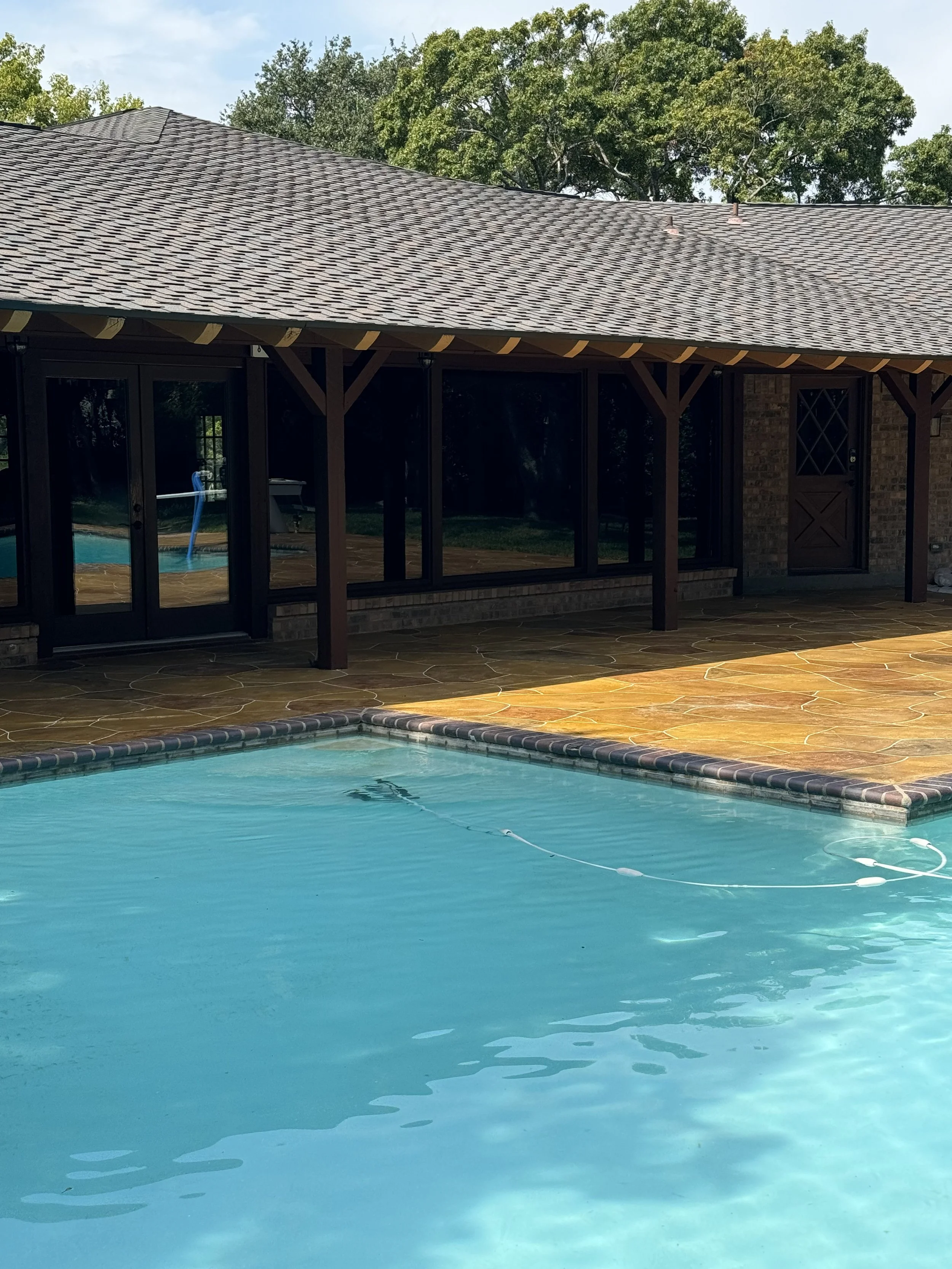 Backyard swimming pool with clear blue water, with a pool cleaning or filtration device floating on the water, surrounded by a stone patio, with a house in the background that has a large screened porch and a windowed door, and trees with green leaves behind the house.