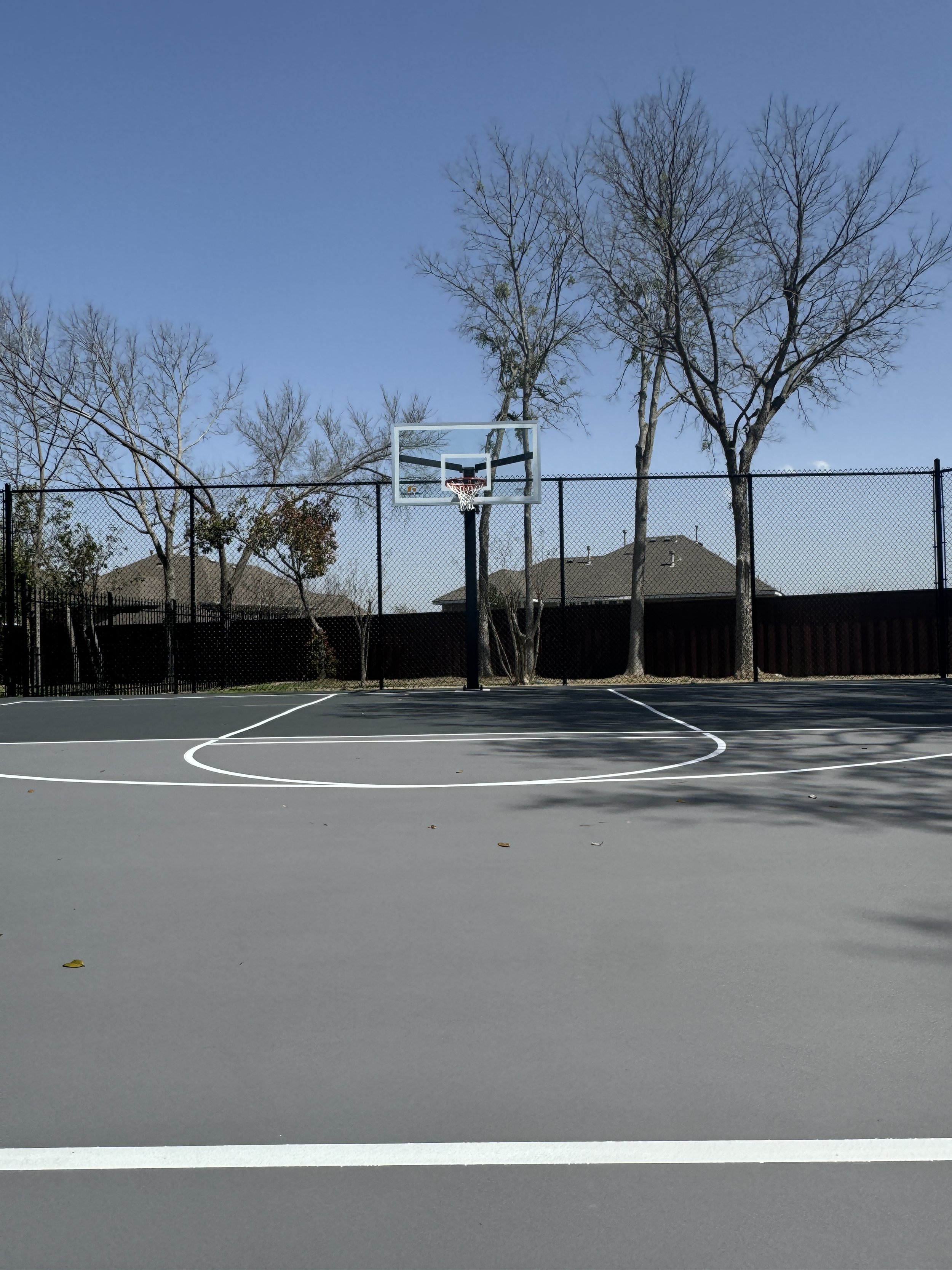 An outdoor basketball court with a hoop, surrounded by a fence, with leafless trees and rooftops in the background under a clear blue sky.