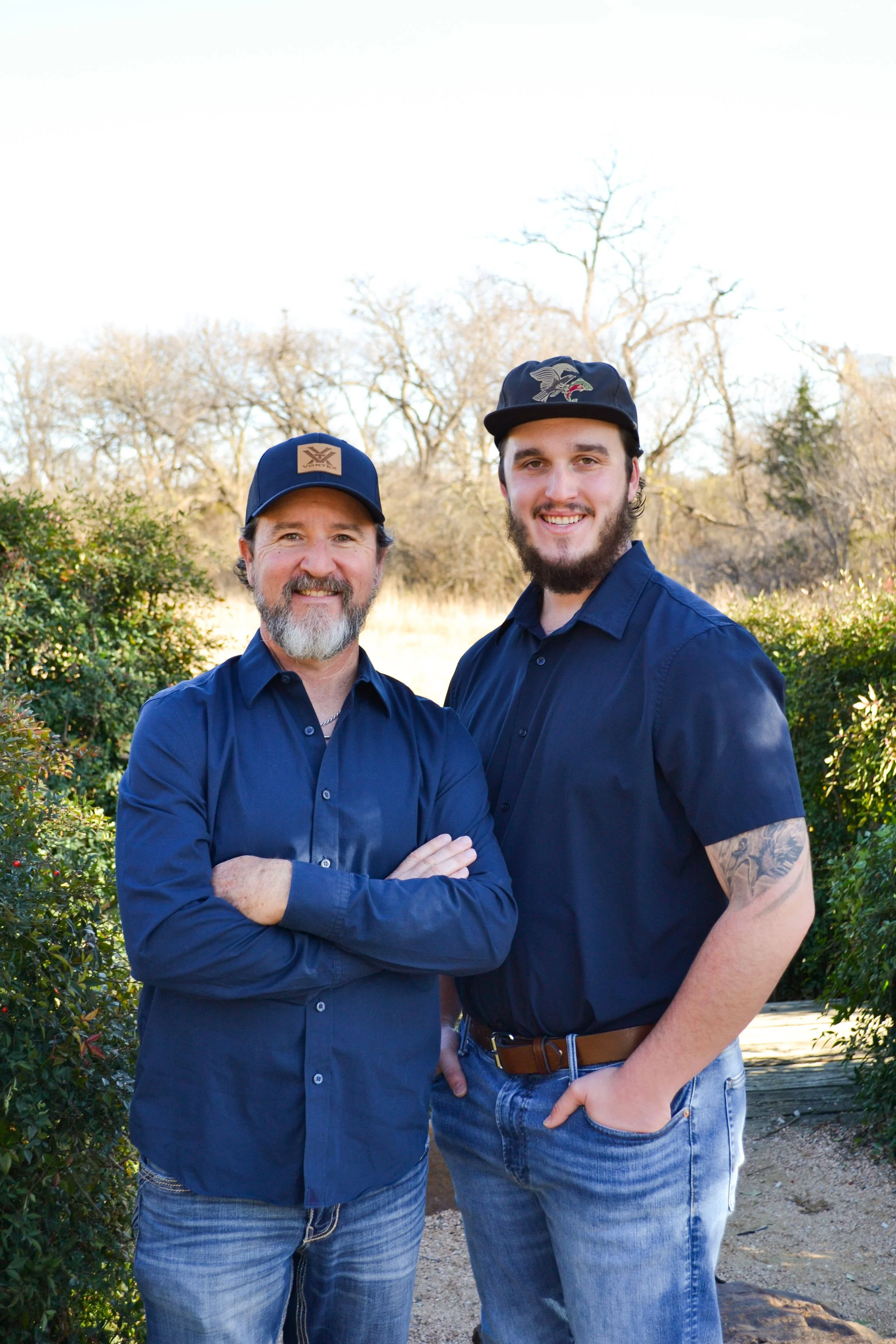Two men smiling outdoors, wearing dark blue shirts and caps, standing in front of leafless trees and green bushes.