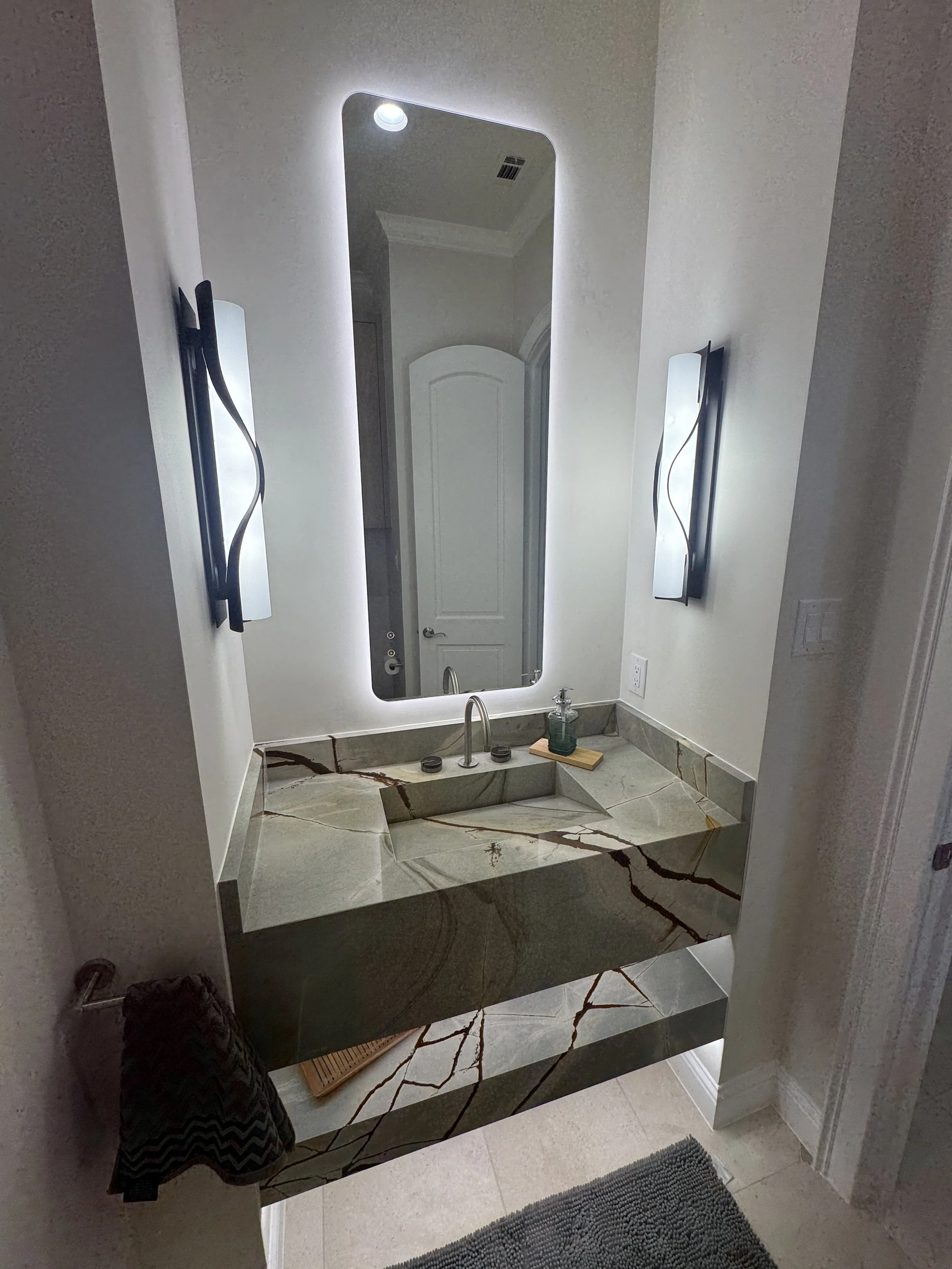 Modern bathroom with marble countertop vanity, large mirror, and wall-mounted lights, beige walls, and entry door reflected in the mirror.