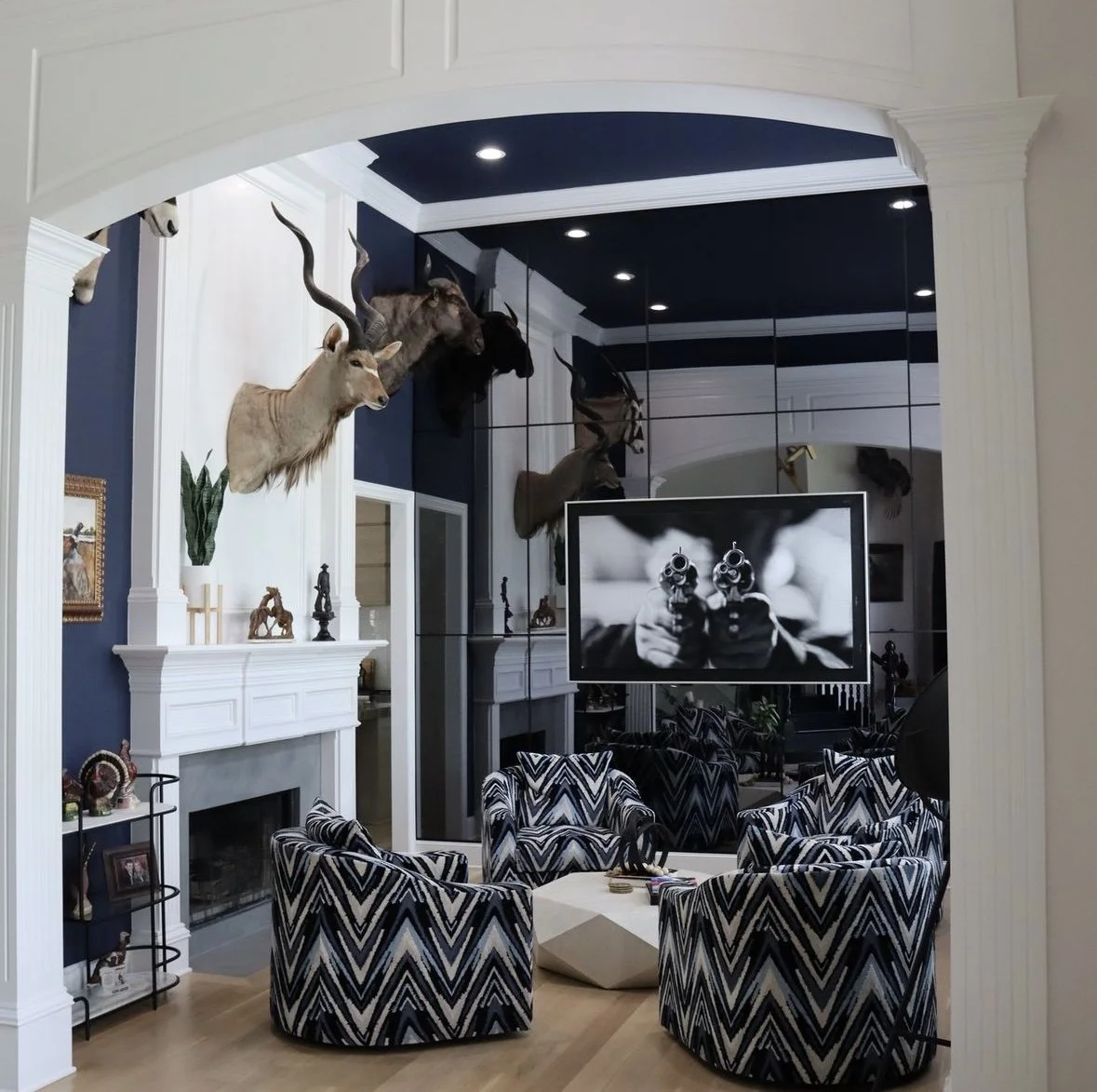 Living room with taxidermy animal heads on the wall, a sitting area with four patterned chairs around a geometric coffee table, a fireplace, and a large flat-screen TV displaying a black-and-white image of a person aiming guns.