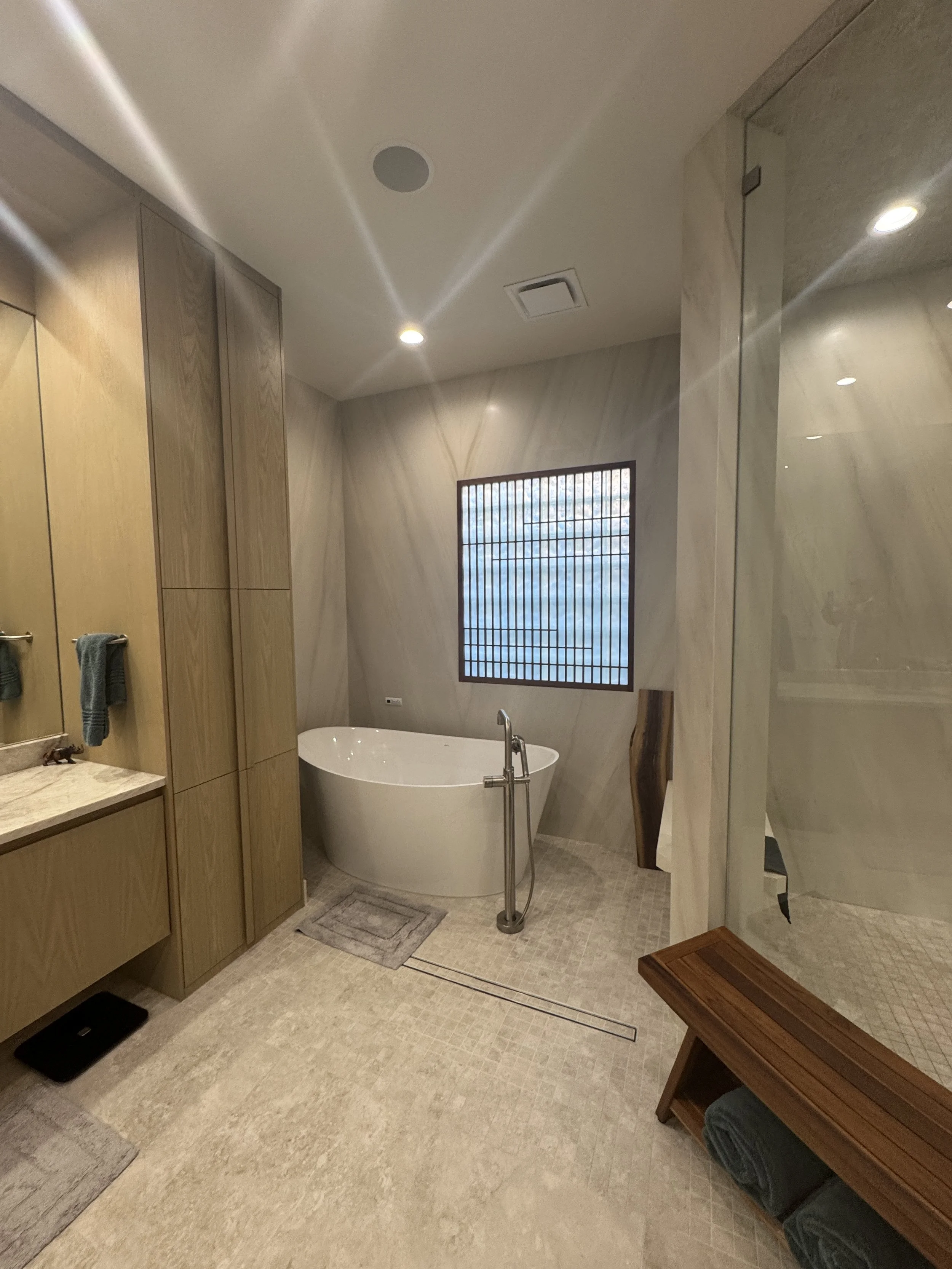 A modern bathroom with a freestanding bathtub near a window with wooden grid design, a wooden bench, and a tiled floor. Walls are light-colored marble, with a mirrored wall and wooden cabinets.