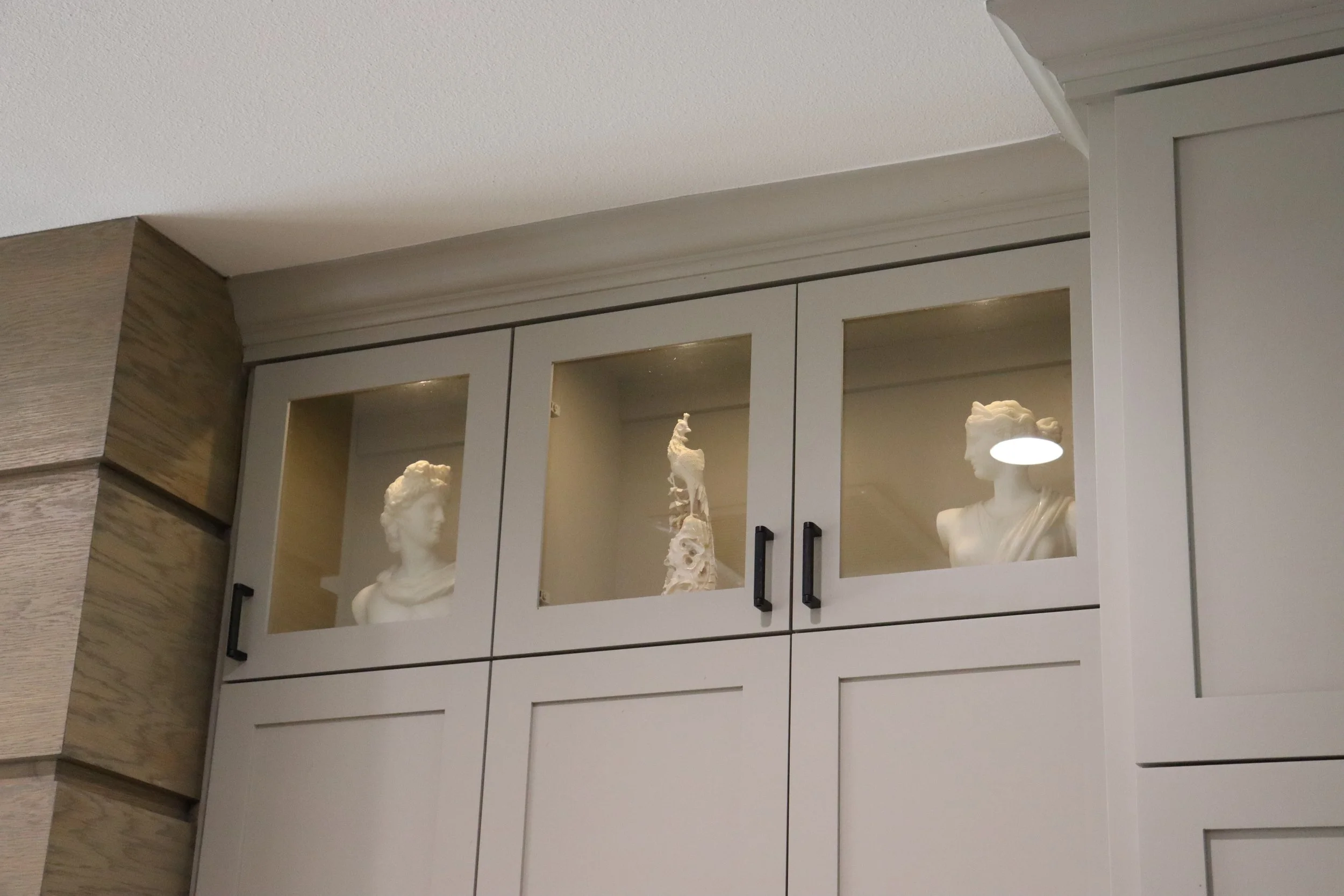 A display cabinet with three glass doors containing white sculpture busts of women and a skeletal model, mounted on a wall with wood paneling in a room with a white textured ceiling.
