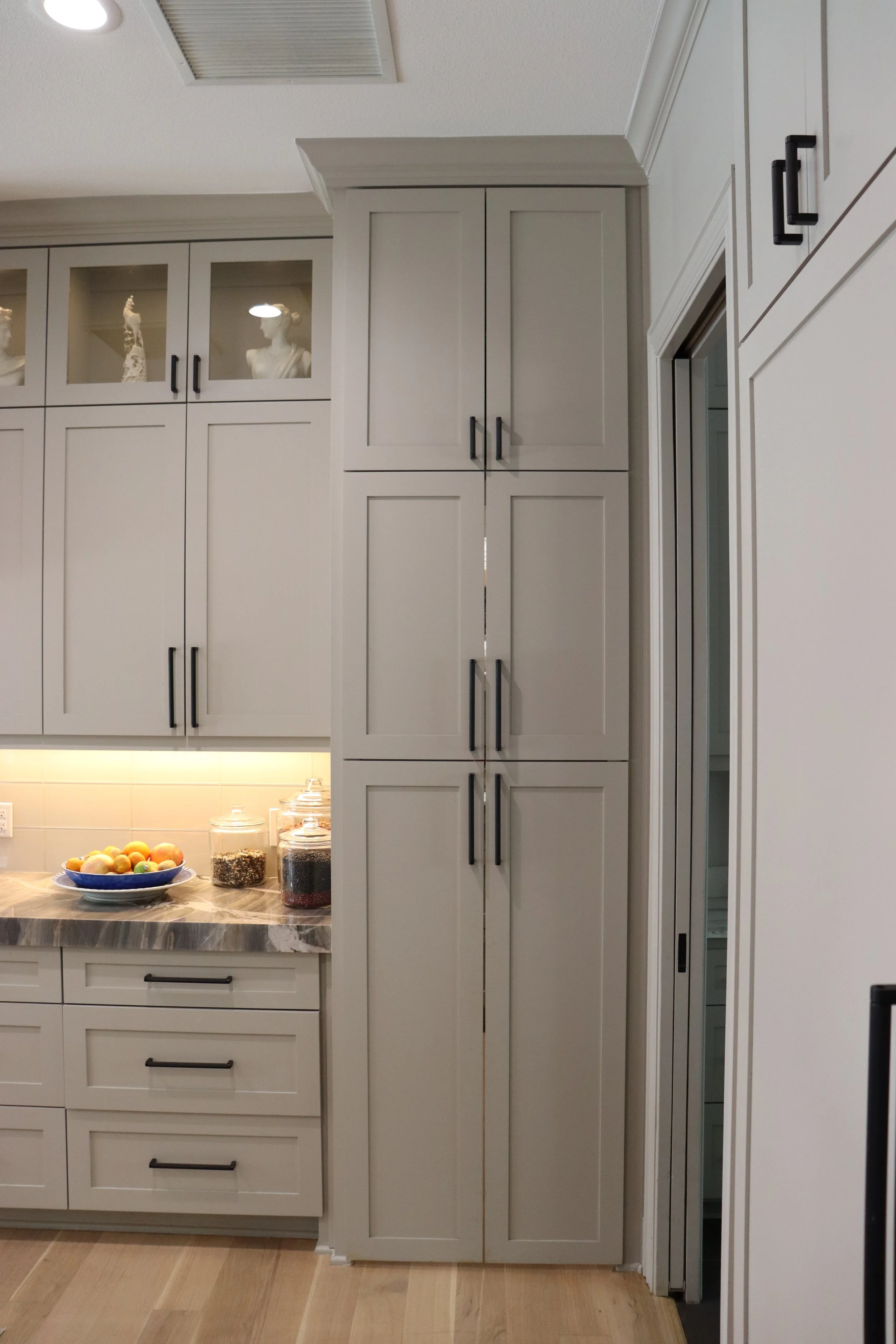 A kitchen with white cabinets, a marble countertop, and a bowl of various fruits on the counter. There are decorative bust sculptures and glass jars with food items on the cabinets. The floor is wooden.