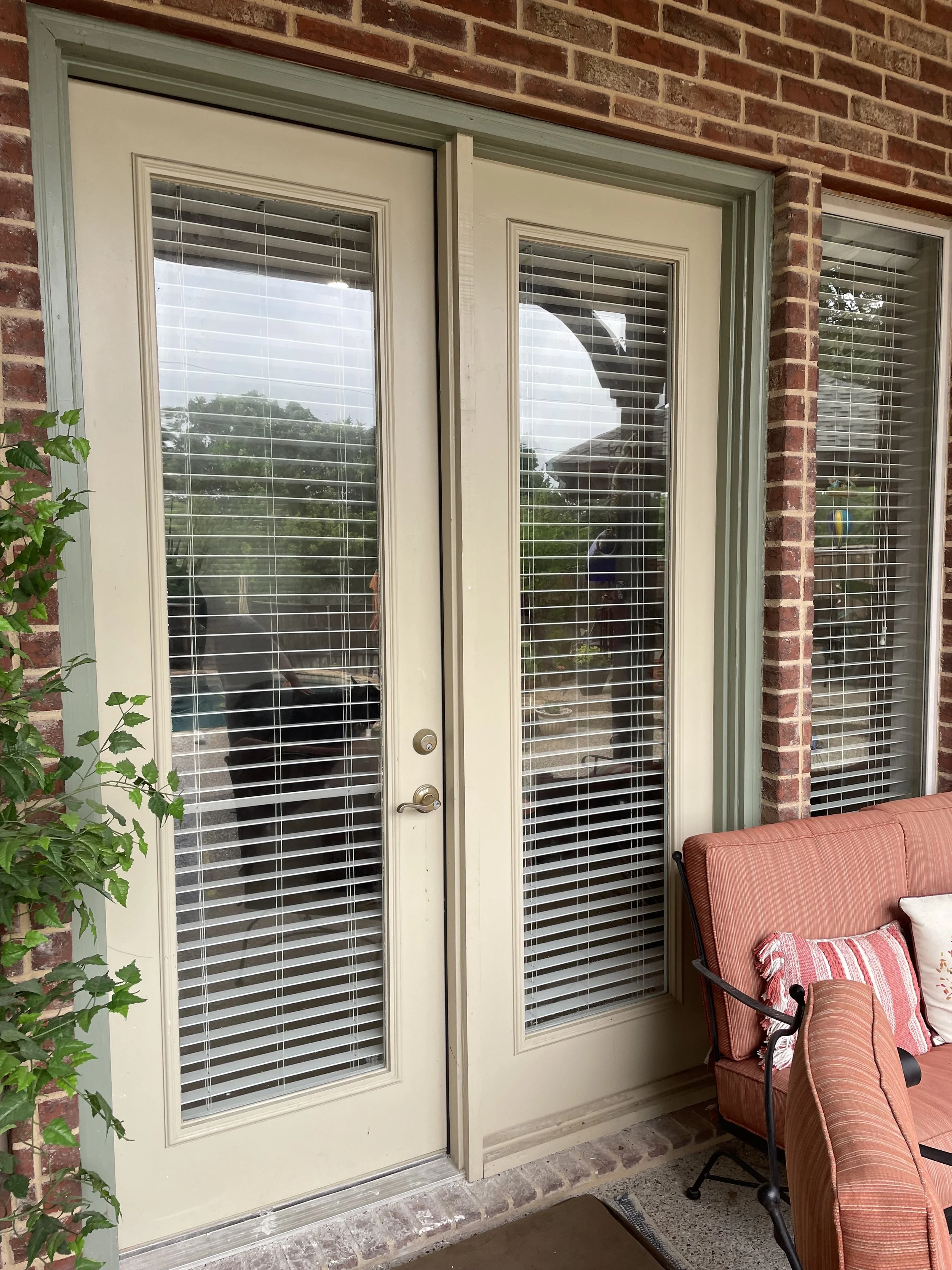 Outside patio door with glass panels and white blinds, a red cushioned chair with a striped pillow is next to the door, and green vines growing on the left side of the image.