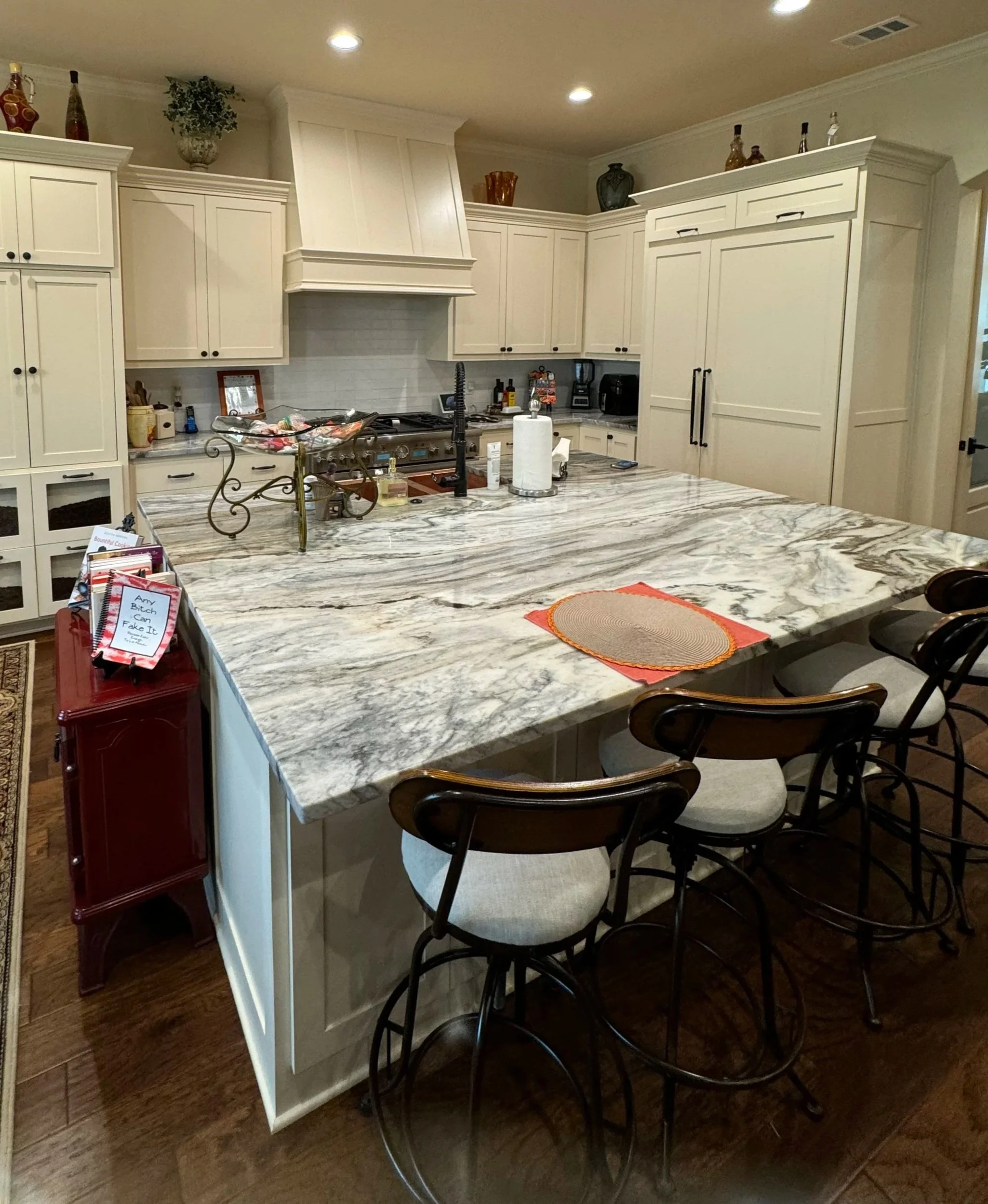 Kitchen with marble island countertop, cream cabinets, and barstools, decorated with vases and bottles on top of cabinets, and various kitchen appliances and items on the counters, including a paper towel roll and snacks.