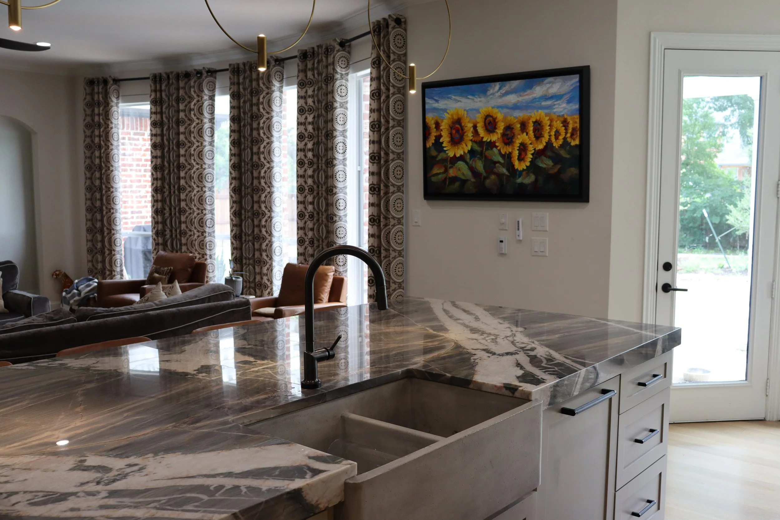 A modern kitchen with a marble countertop, black sink, white cabinets, and a view into a living room with large windows, patterned curtains, a painting of sunflowers on the wall, and outdoor greenery.