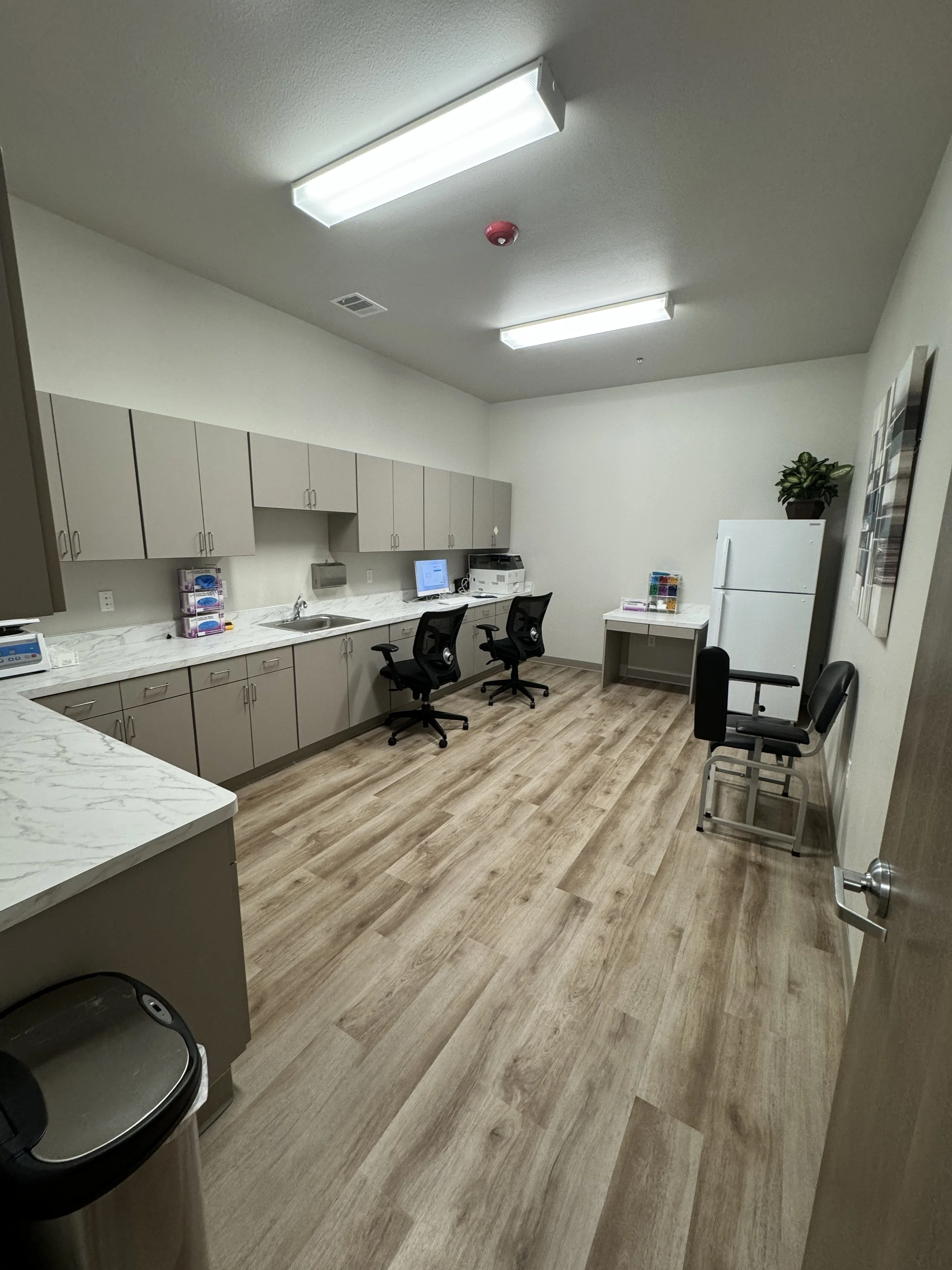 A clean, well-lit office or medical room with cabinetry, chairs, a computer, and a small refrigerator with a potted plant on top.