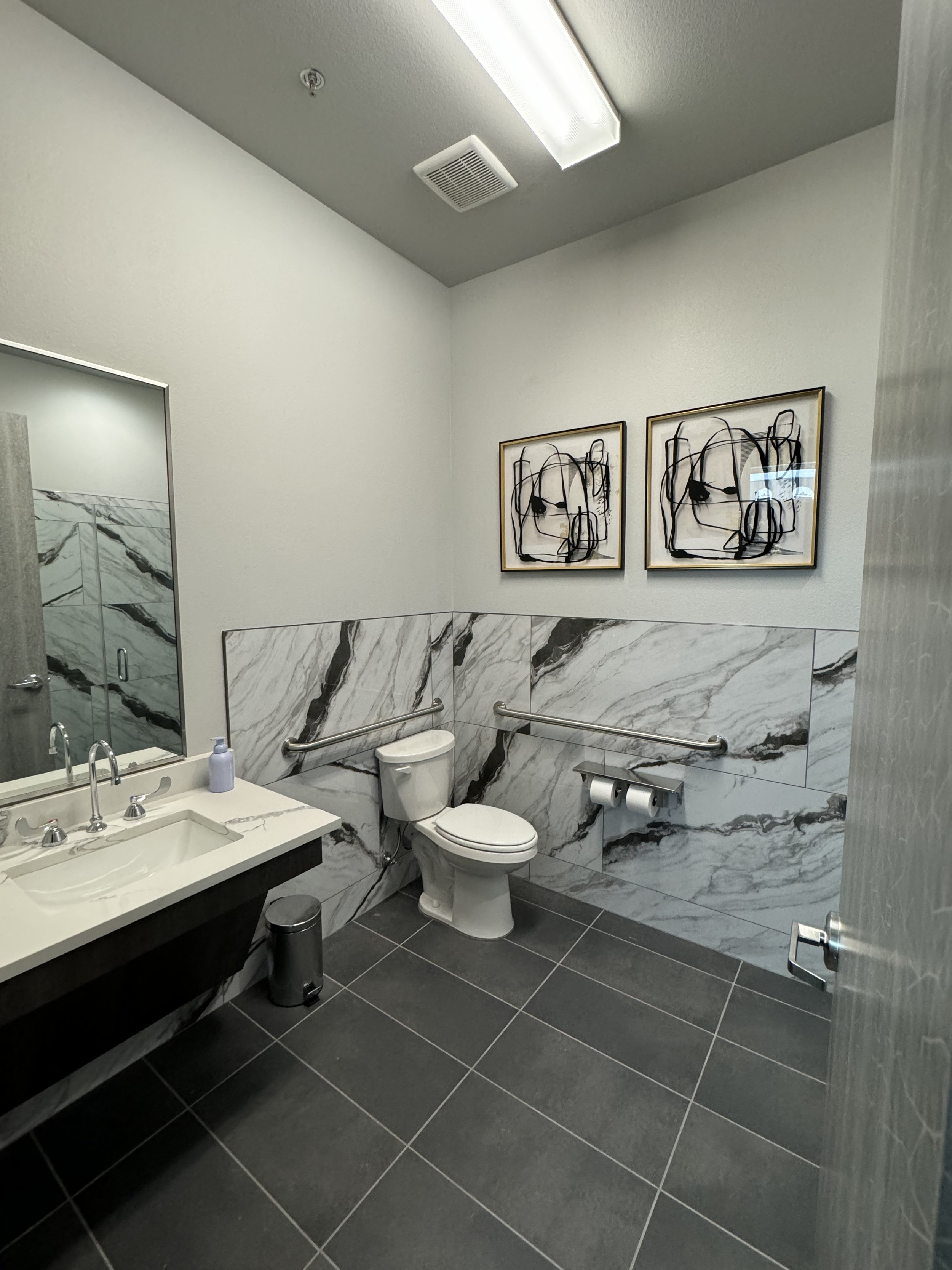 Public restroom with marble walls and flooring, a mirror above a white sink with a soap dispenser, a toilet with a grab bar, and two framed abstract art pieces on the wall.