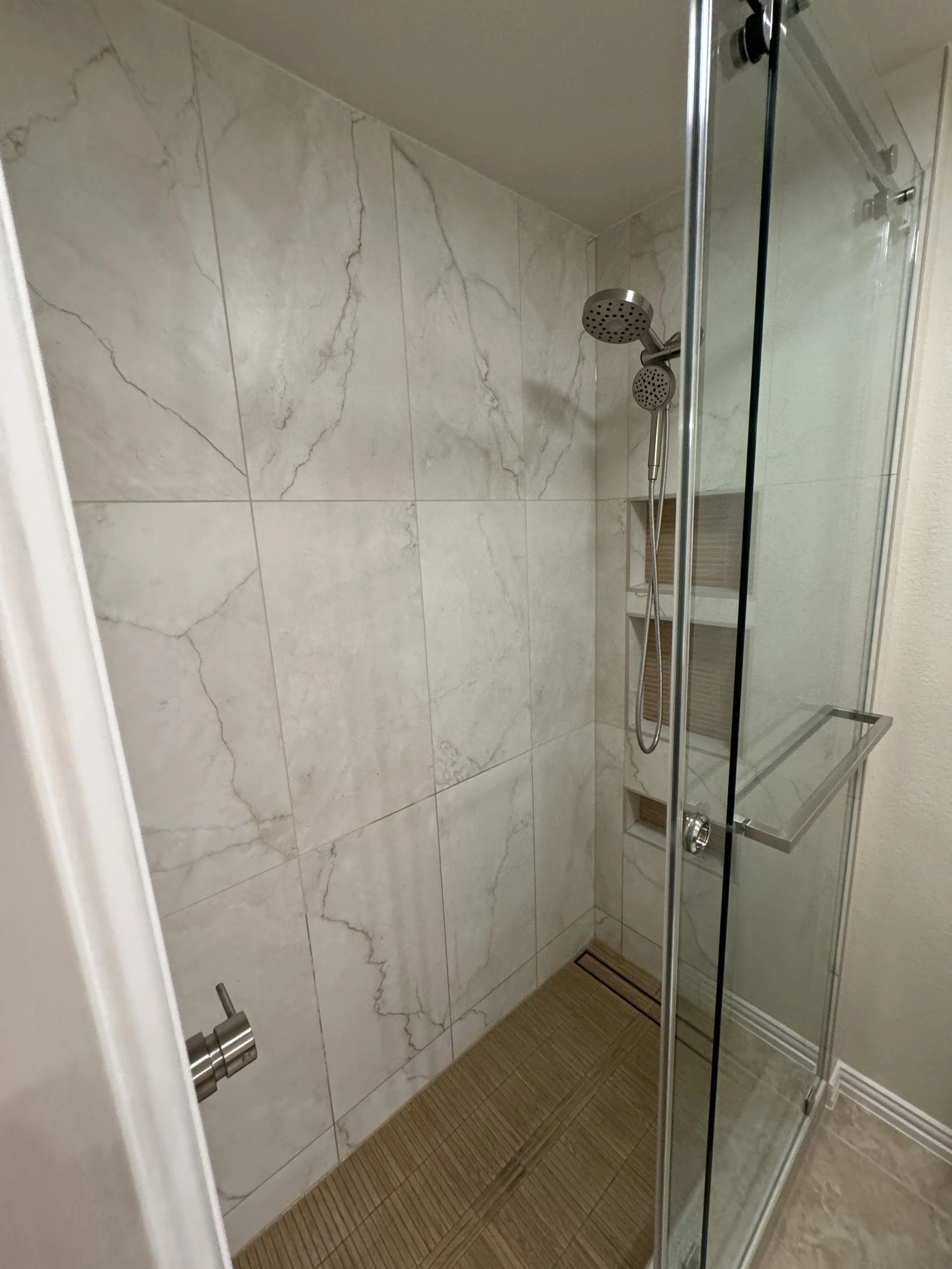 A modern shower with beige marble tiled walls, a glass door, a handheld shower head, and a wooden shower mat on the floor.