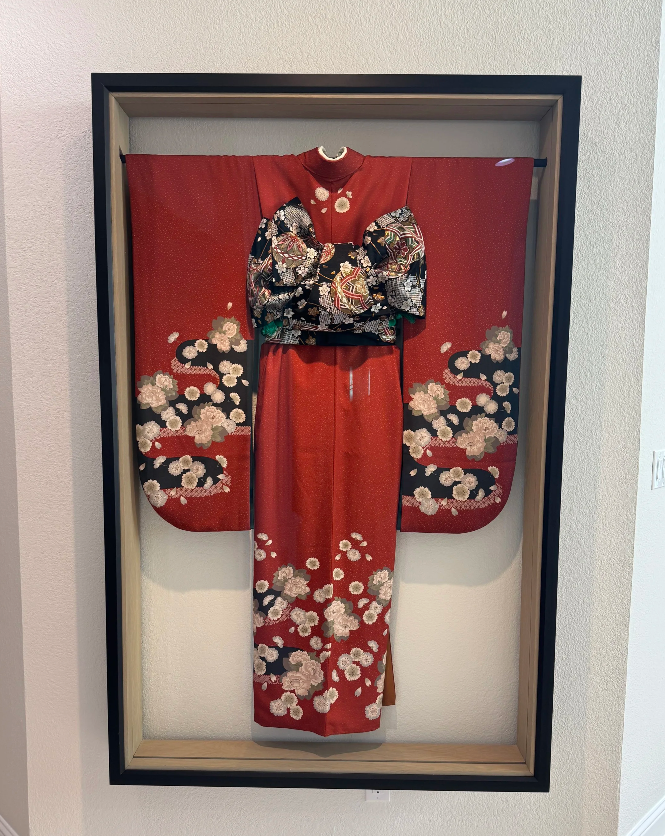 A traditional Japanese kimono on display, featuring a red base with floral designs, including pink and white flowers, displayed in a shadow box frame.