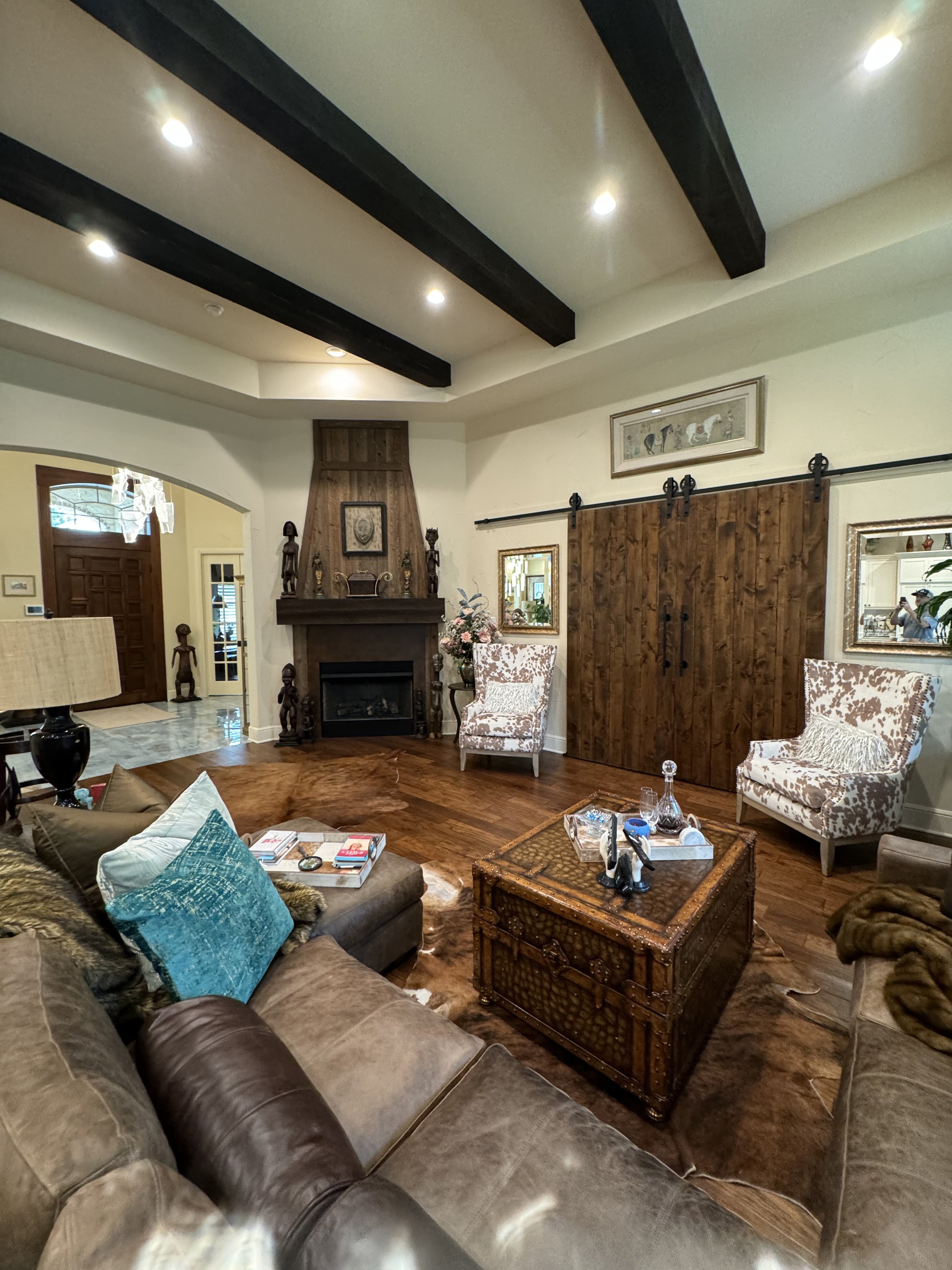 A living room with a large sofa, armchair, wooden coffee table, fireplace, decorative items, framed art, and a barn door, illuminated by ceiling lights.