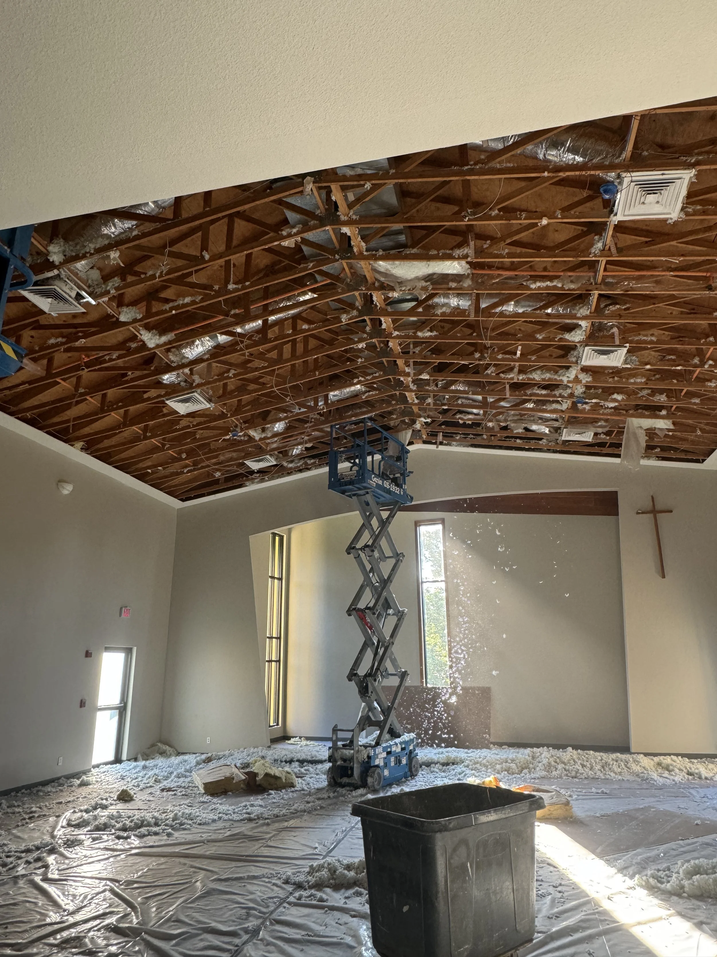 Construction worker is replacing a ceiling in a large room with high ceilings, with insulation and air vents visible. The room has windows and is bright with natural light.