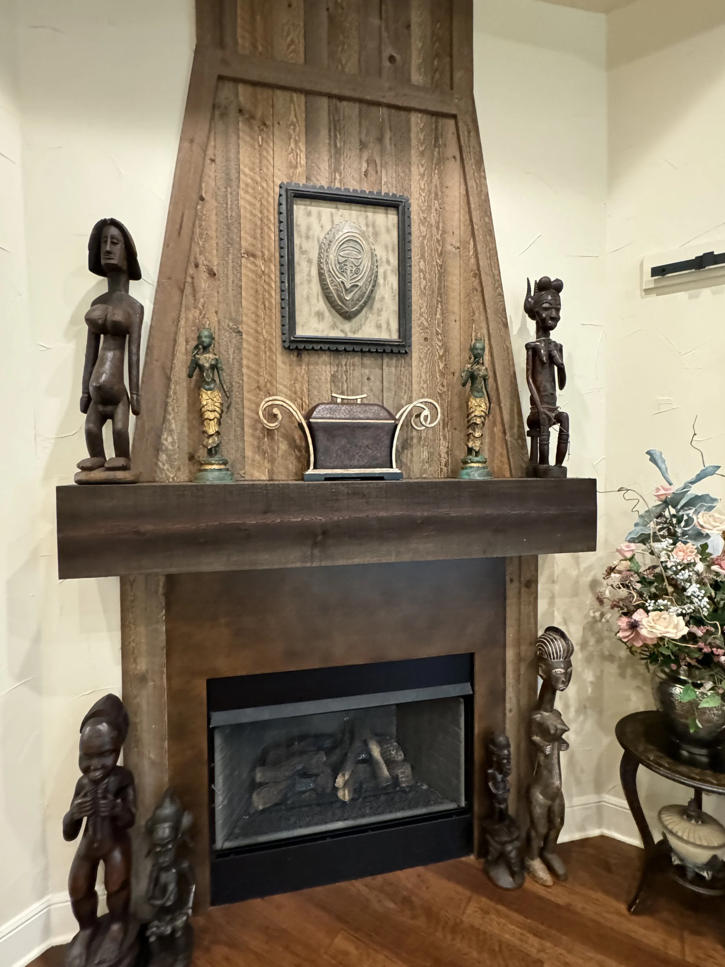Decorative fireplace with tribal African statues on either side, a framed artwork above the mantle, and a side table with flowers to the right.