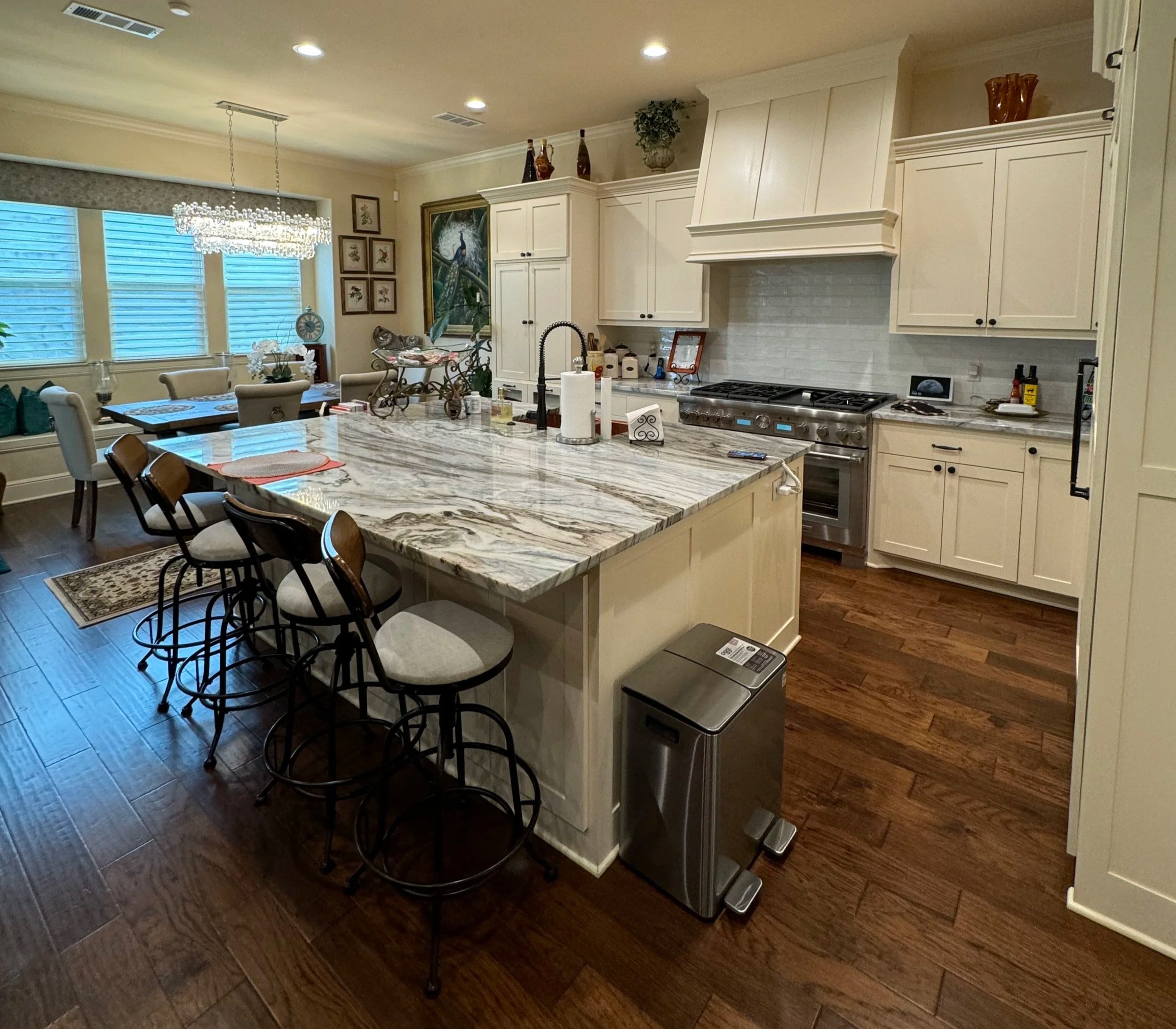 A modern kitchen with a large marble island, white cabinets, and a stainless steel oven. There are chairs around the island, and decorative items on top, including paper towel rolls and small objects. The room has hardwood floors, framed artwork on the walls, a dining area with a chandelier, and a large window with blinds.