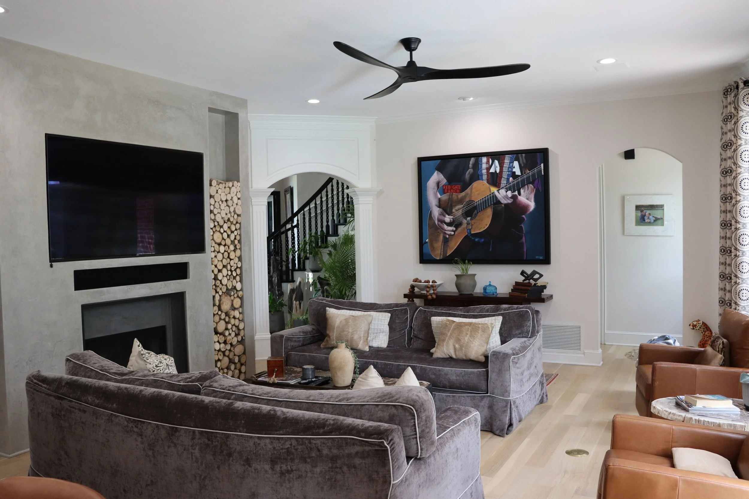 Living room with gray velvet sofas, a wall-mounted flat-screen TV, a ceiling fan, a staircase, and a large framed picture of a person playing guitar.