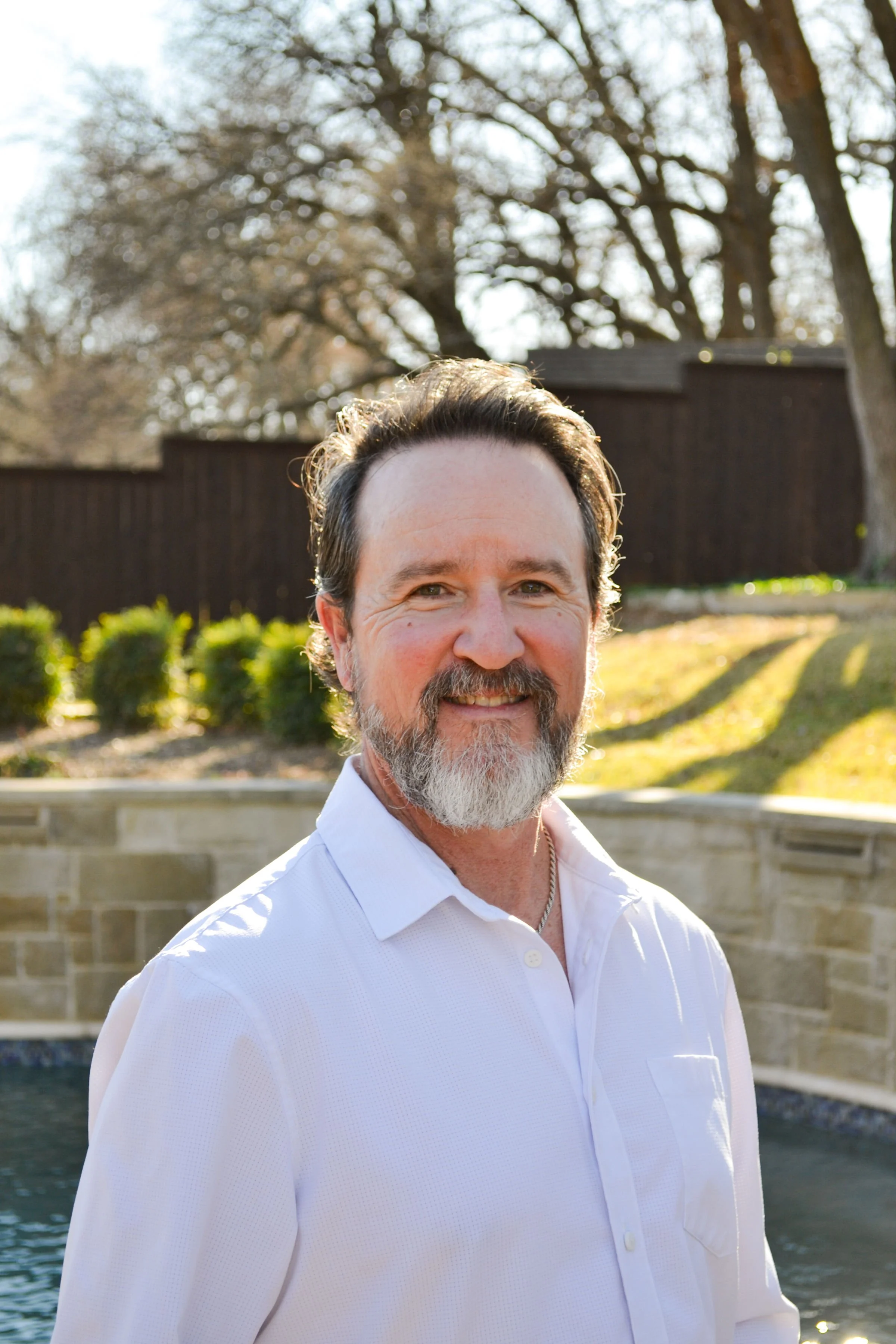 A middle-aged man with a beard and mustache, smiling outdoors on a sunny day. He is wearing a white button-up shirt, standing in front of a pond and a landscaped yard with trees, bushes, and a wooden fence in background.