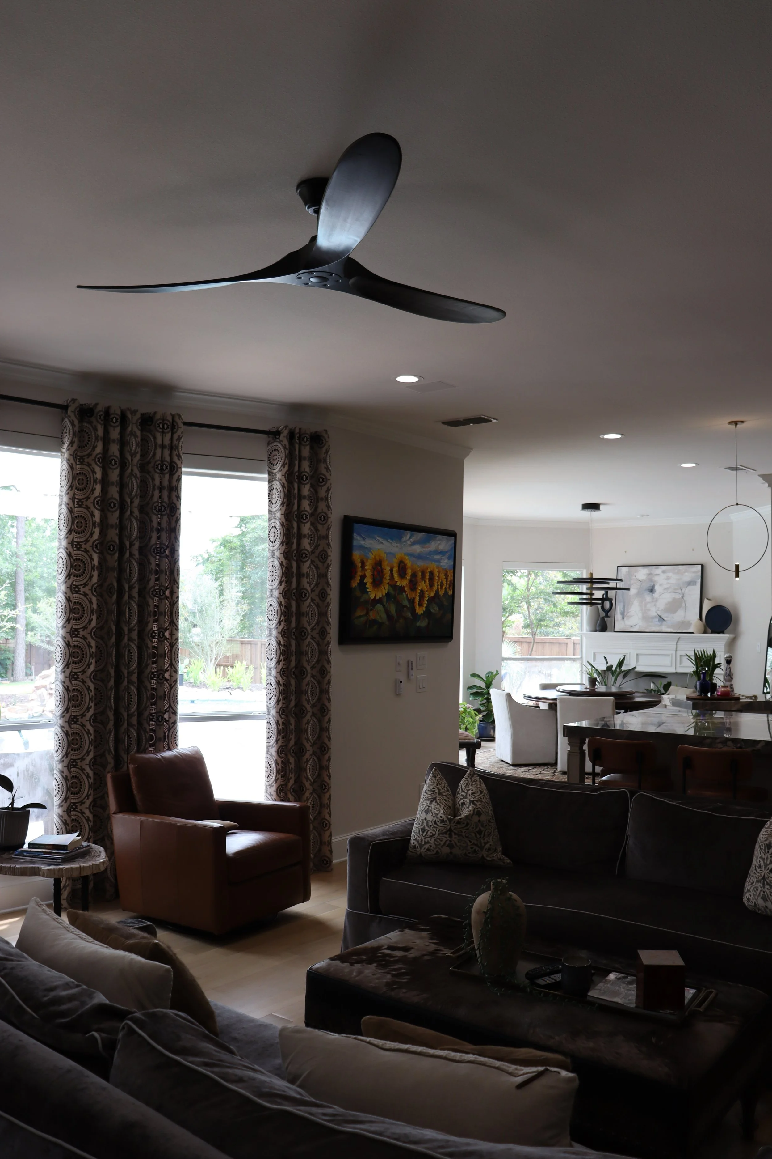 View of a living room with a ceiling fan, large windows with patterned curtains, a painting of sunflowers on the wall, and various furniture including chairs and sofas with decorative pillows.