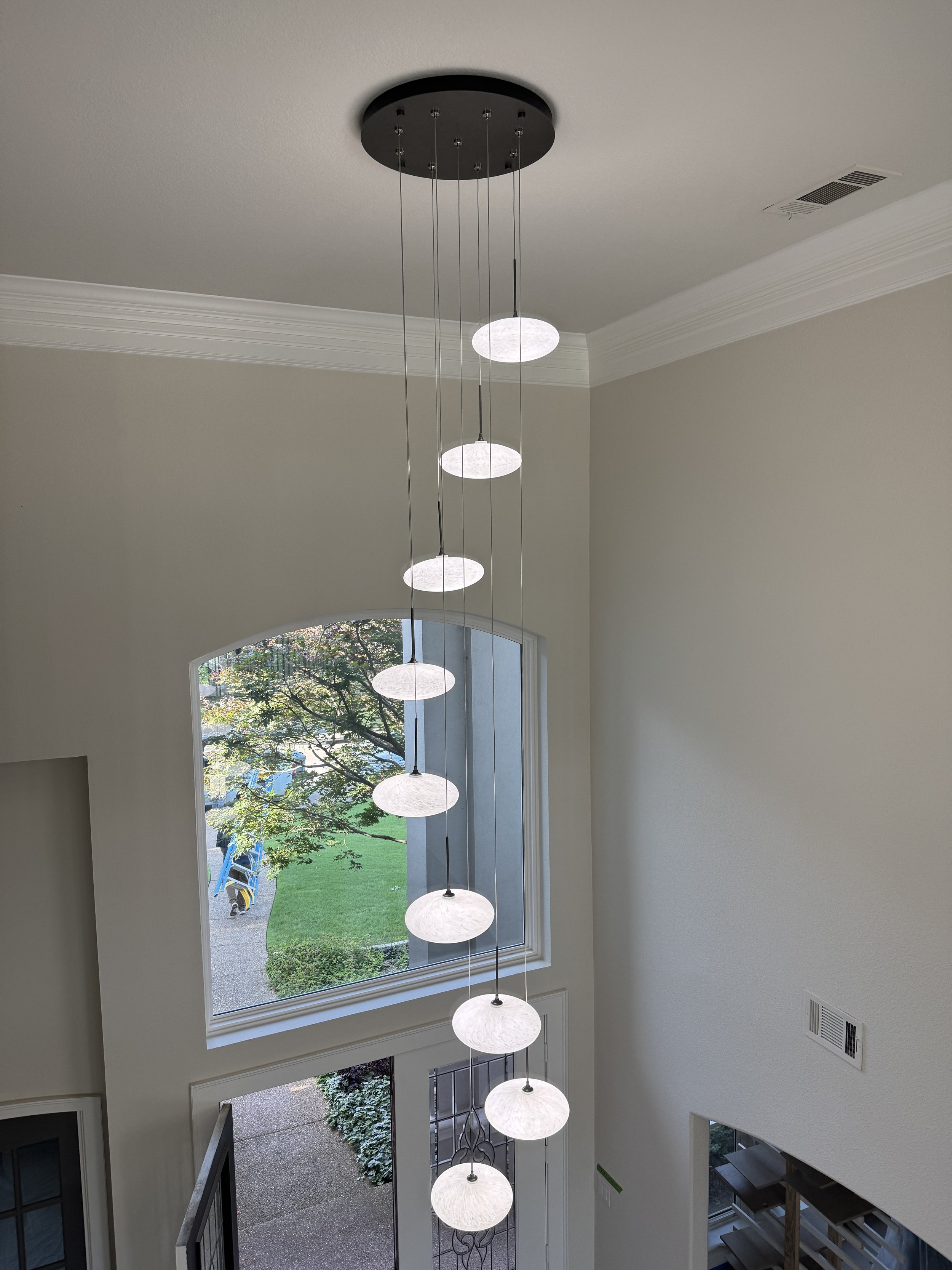Interior view of a spacious room with a tall ceiling, featuring a modern hanging light fixture with multiple round white lamps, large rectangular window showing a garden with trees and a blue object outside, and a patio area with greenery.