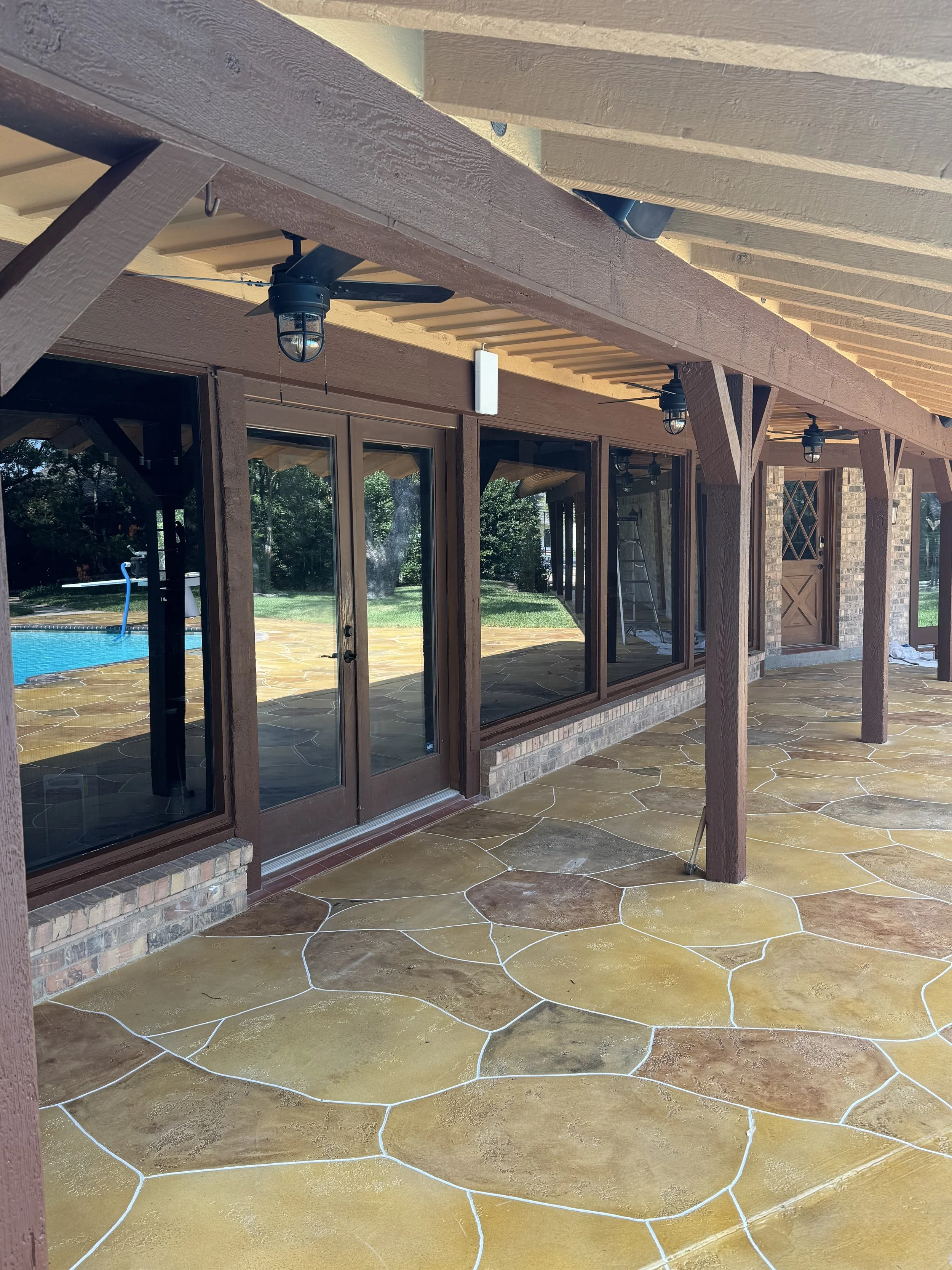 A view of a patio area with a yellow stained concrete floor with white grout lines, wooden support beams, and a screened-in porch with hanging light fixtures. There are glass sliding doors leading inside, and trees and backyard grass are visible outside.
