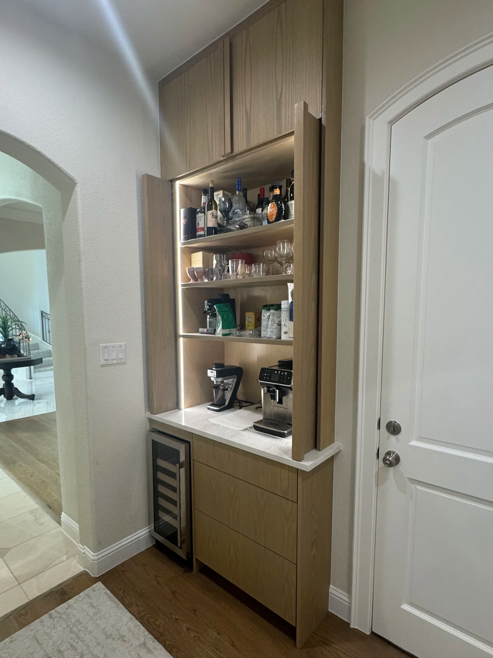 A home bar with wooden cabinets, shelves with liquor bottles and glassware, a coffee machine, and a mini refrigerator, placed against a wall near a white door.