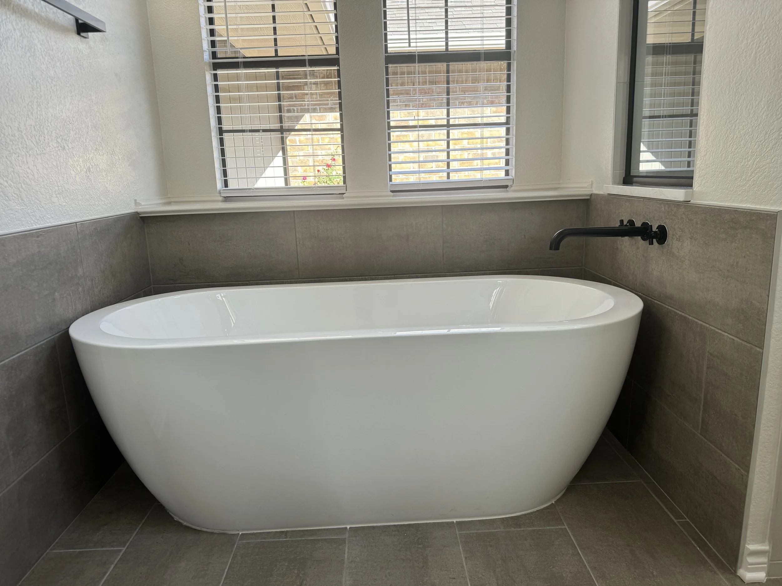A modern bathtub in a bathroom with neutral-colored tiles and white walls, with three large windows with white blinds letting in natural light.