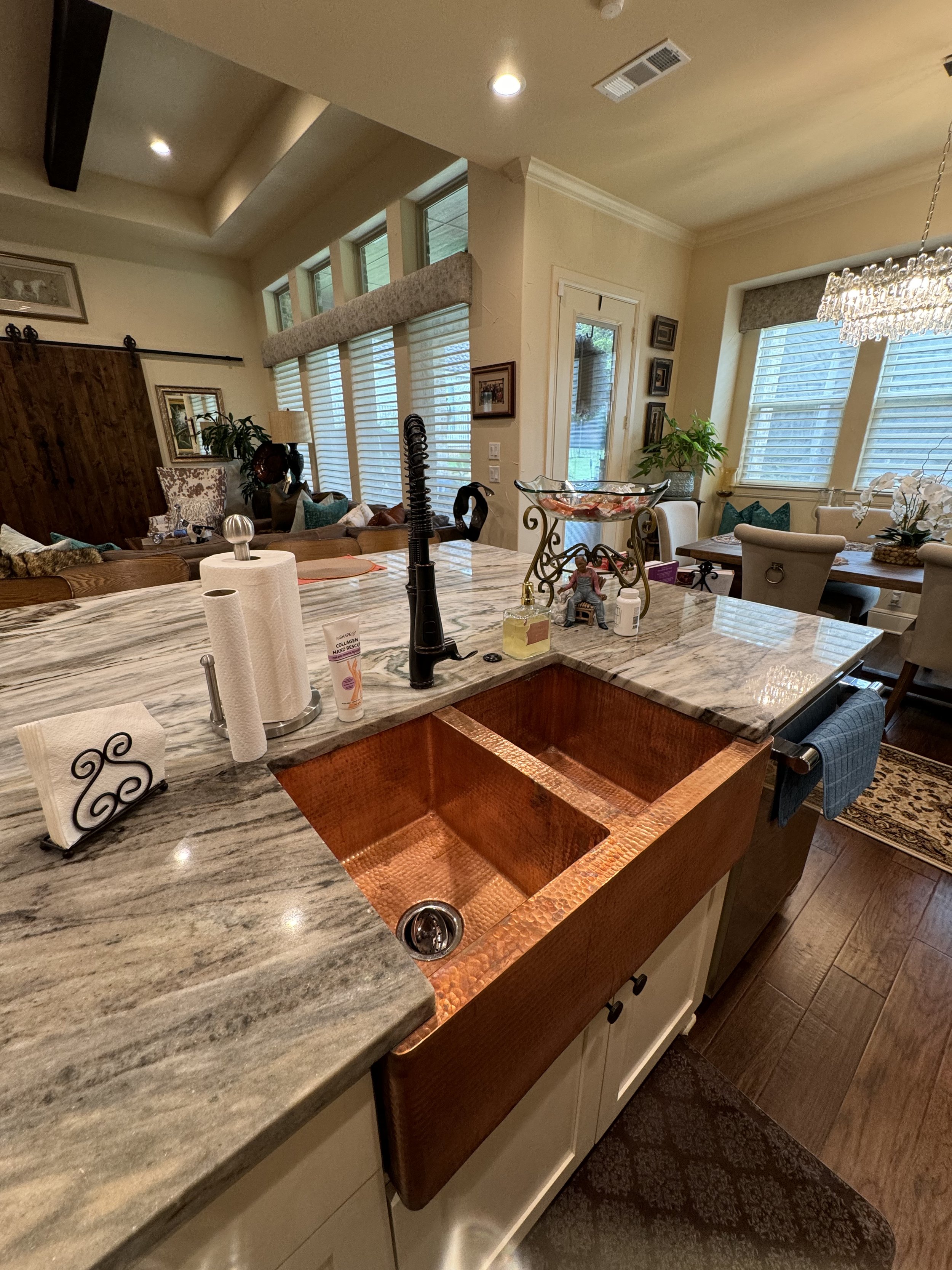 The photo depicts a kitchen with a marble countertop, a copper sink, paper towels, and various small items including a bottle of hand soap and a figurine. In the background, there are cozy seating areas with cushions, plants, and large windows with blinds. The ceiling features recessed lighting and a chandelier.