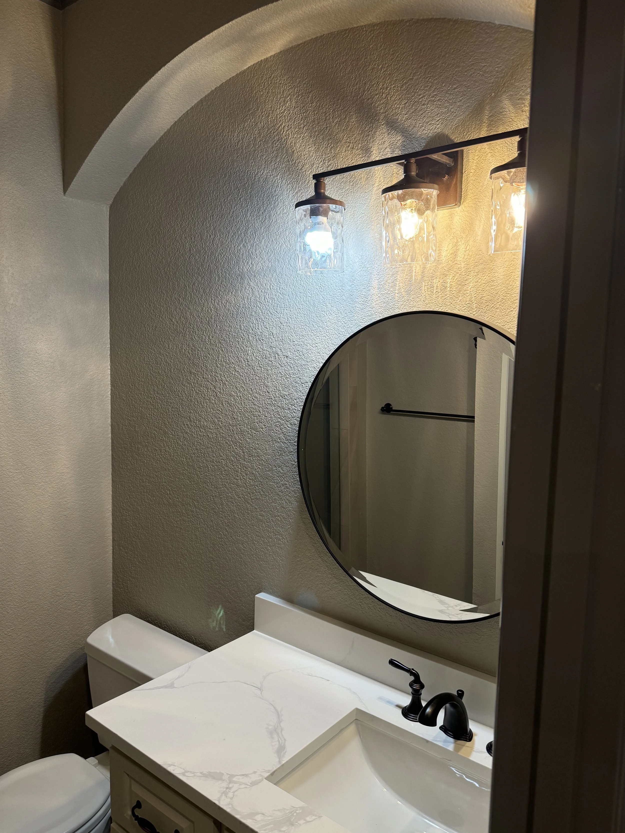 Bathroom with a white marble vanity, black faucet, round mirror, wall-mounted light fixtures with glass shades, beige textured walls, and a toilet.