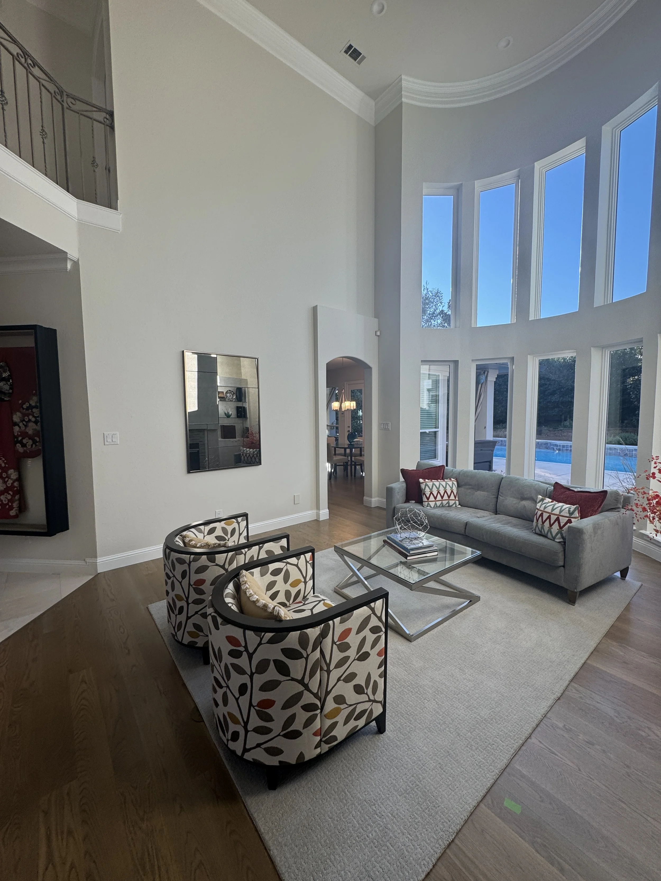A spacious living room with a high vaulted ceiling and large windows showing a backyard pool. The room has a gray couch with patterned pillows, two armchairs with a leaf design, a glass coffee table, and a beige area rug. A dining room is visible through an arched doorway.