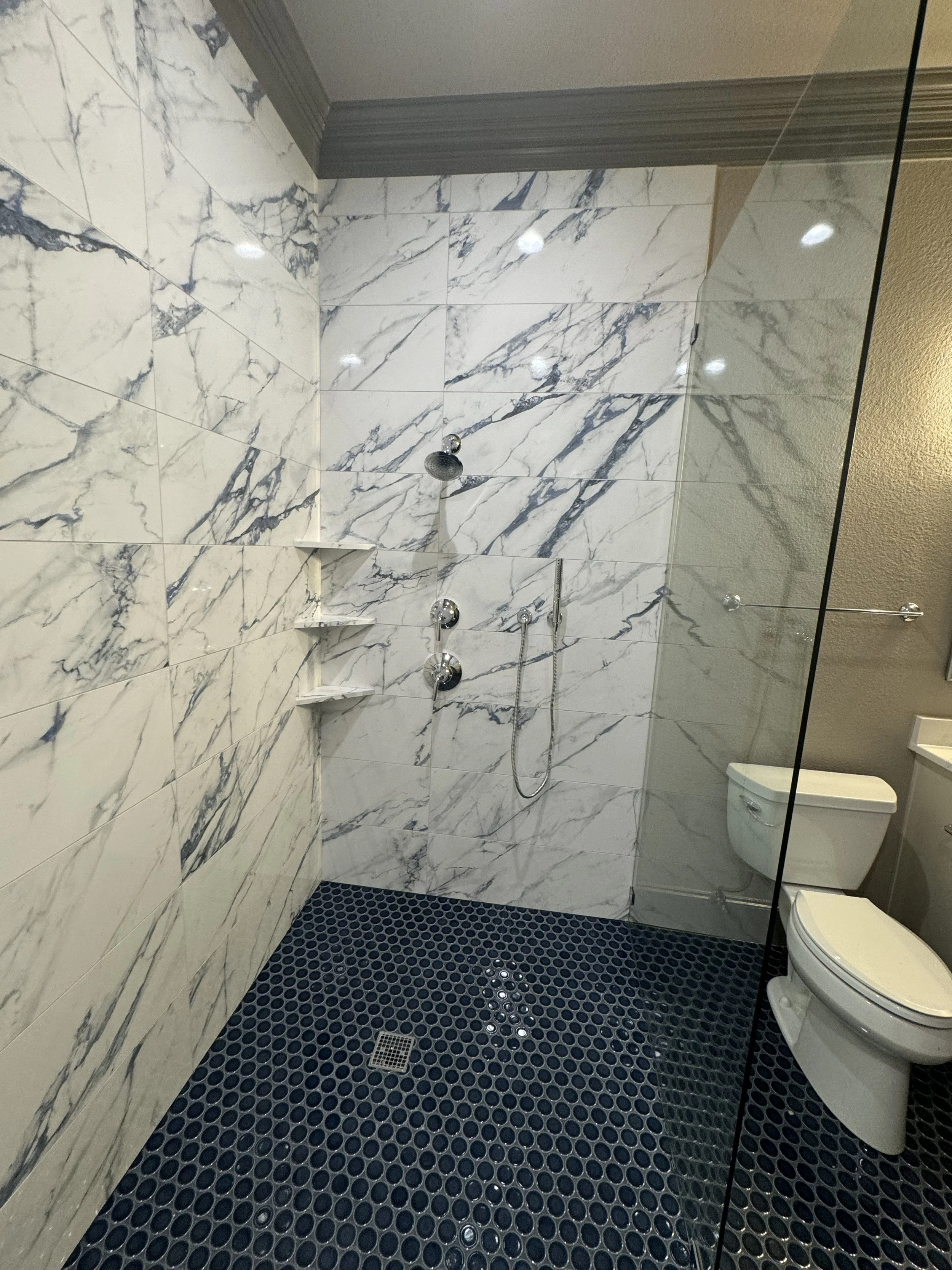 A modern bathroom with marble wall tiles, a shower area with a handheld showerhead, and a black pebble tile floor with a floor drain. There is a white toilet in the corner, a glass shower partition, and some built-in marble shelves in the shower area.
