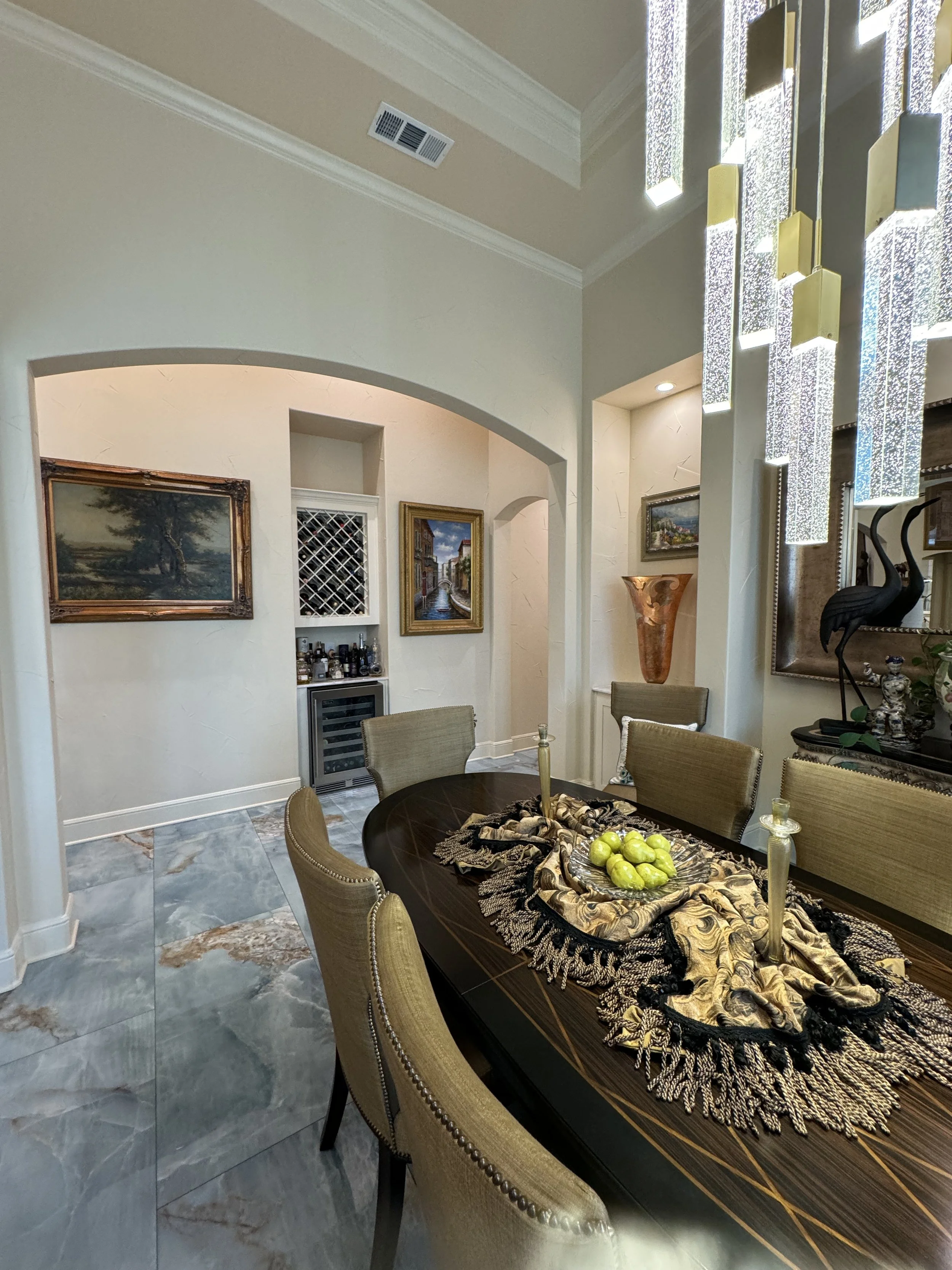 A dining room with a dark wood oval table with a decorative cloth, surrounded by beige upholstered chairs. A centerpiece with green apples is on the table. The room features framed landscape paintings, a tall decorative urn, and black stylized bird sculptures. The space has marble tile flooring, textured white walls, and a modern chandelier hanging from the ceiling. There is a small bar area with liquor bottles and artwork in the background.