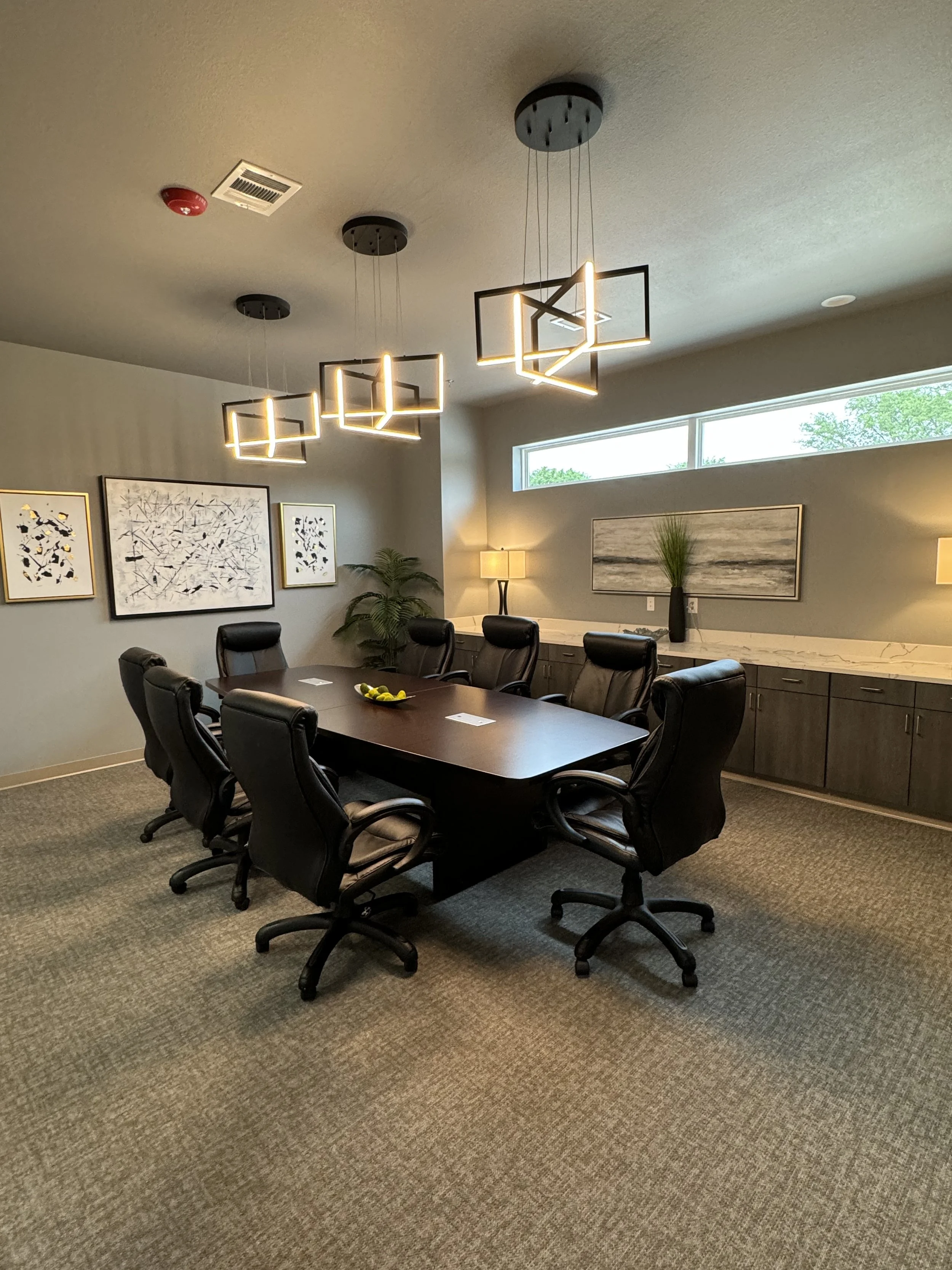 A conference room with a large dark wooden table surrounded by black leather chairs. The room features wall art with abstract designs, a marble sideboard with decorative plants, and modern pendant lighting. There is a narrow horizontal window on the upper wall letting in natural light.