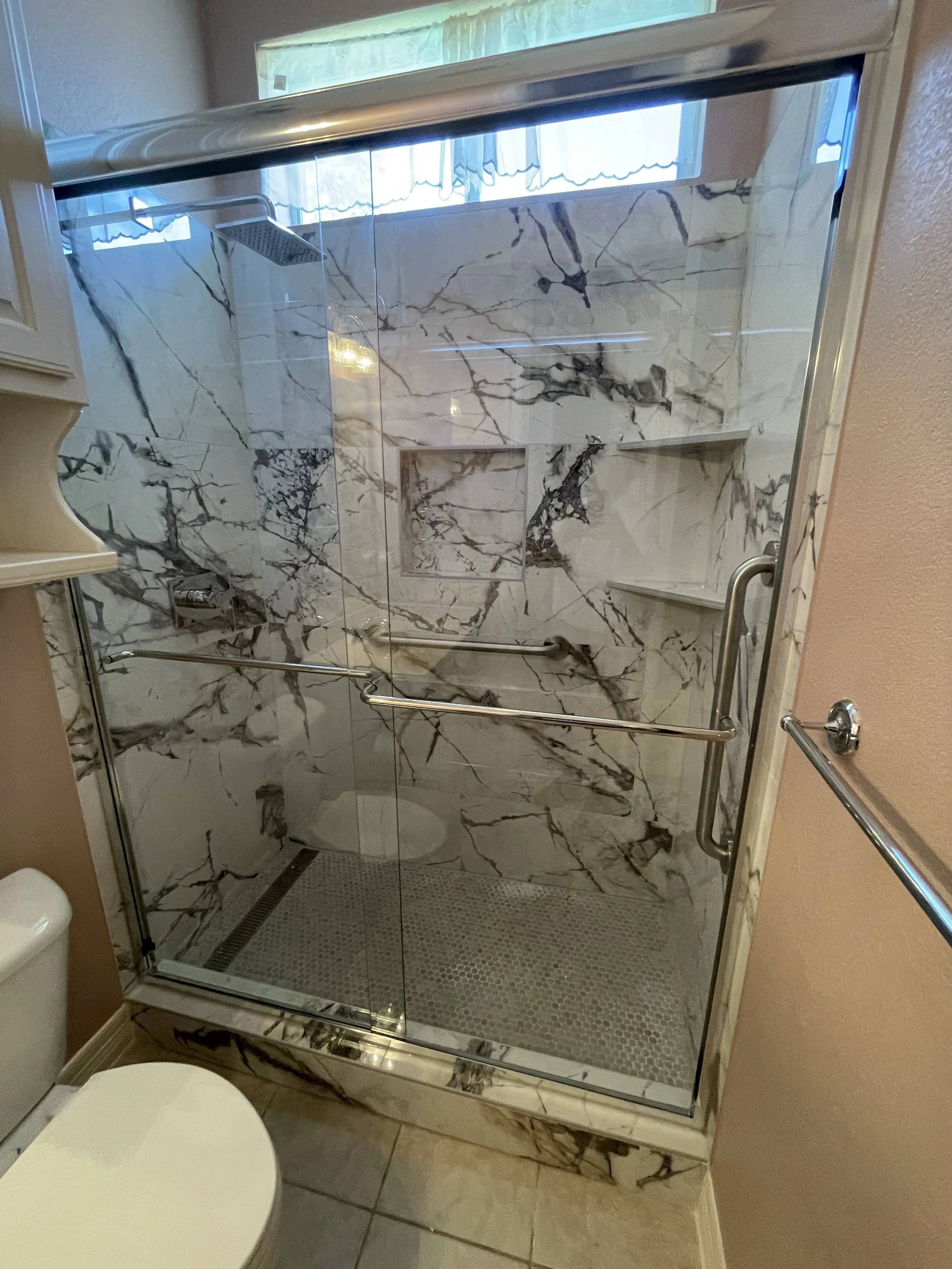 A walk-in shower with marble tiles, a glass door, a built-in shelf, a rainfall showerhead, and a handrail. There is a window with a curtain above the shower.