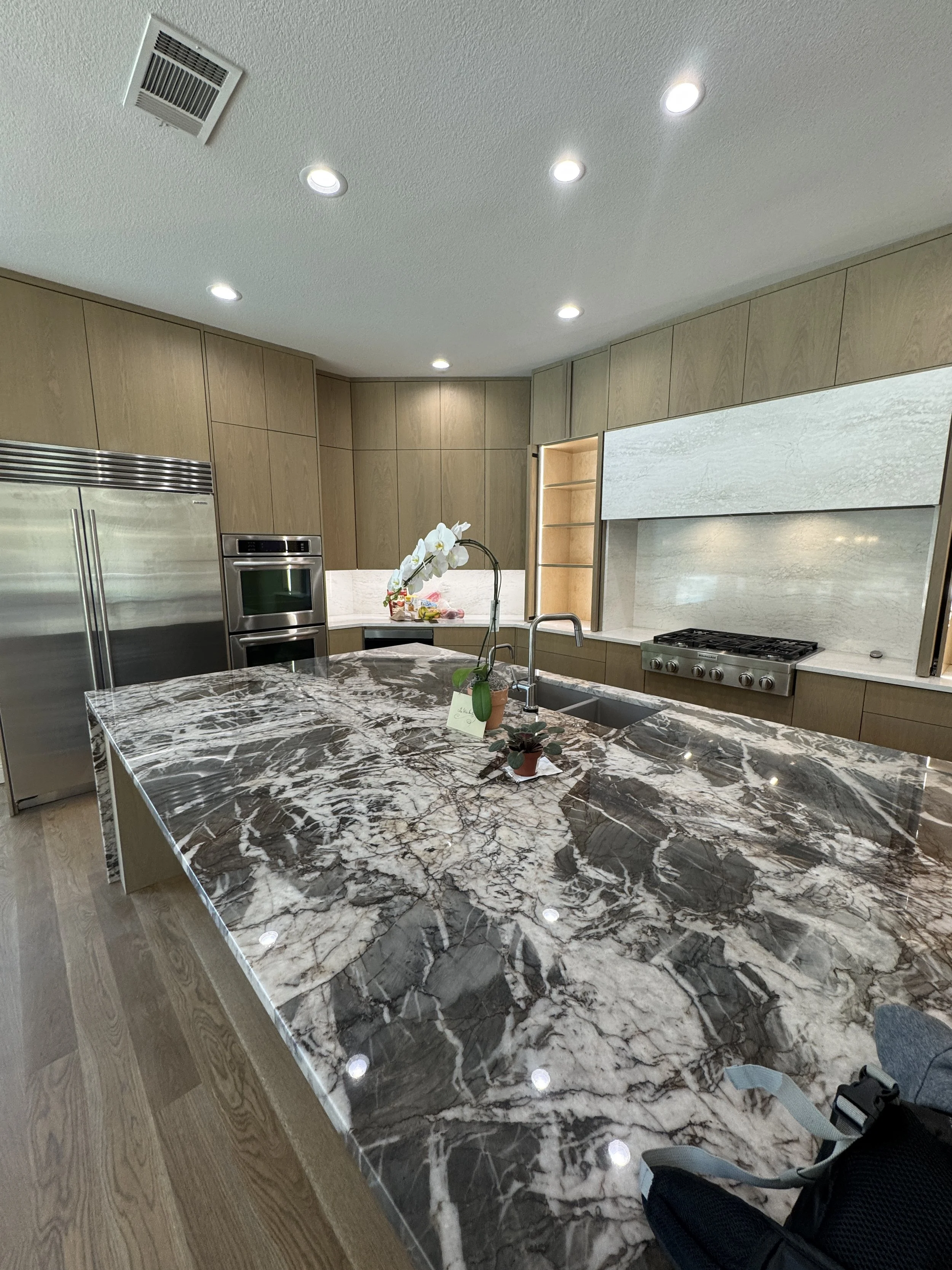 Modern kitchen with wooden cabinets, marble island, stainless steel refrigerator and oven, and a wall with a light-colored marble backsplash. There is an orchid plant on the island and some snacks on the counter.