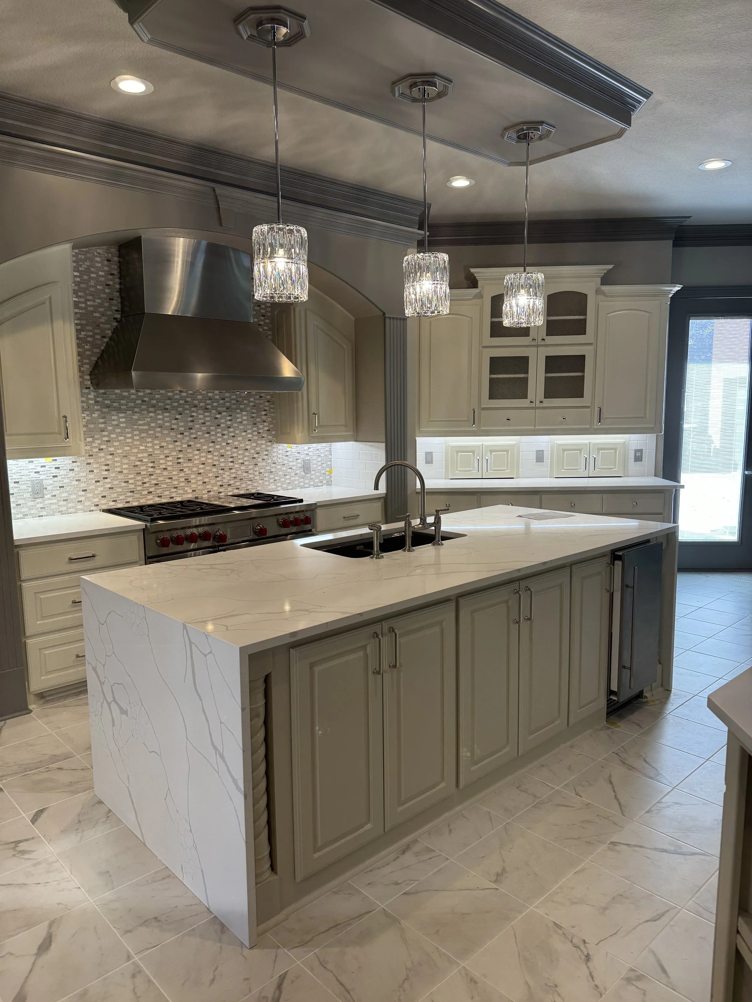 Modern kitchen with a large white marble island, cream-colored cabinetry, a stainless steel range hood, and pendant lights hanging above the island.