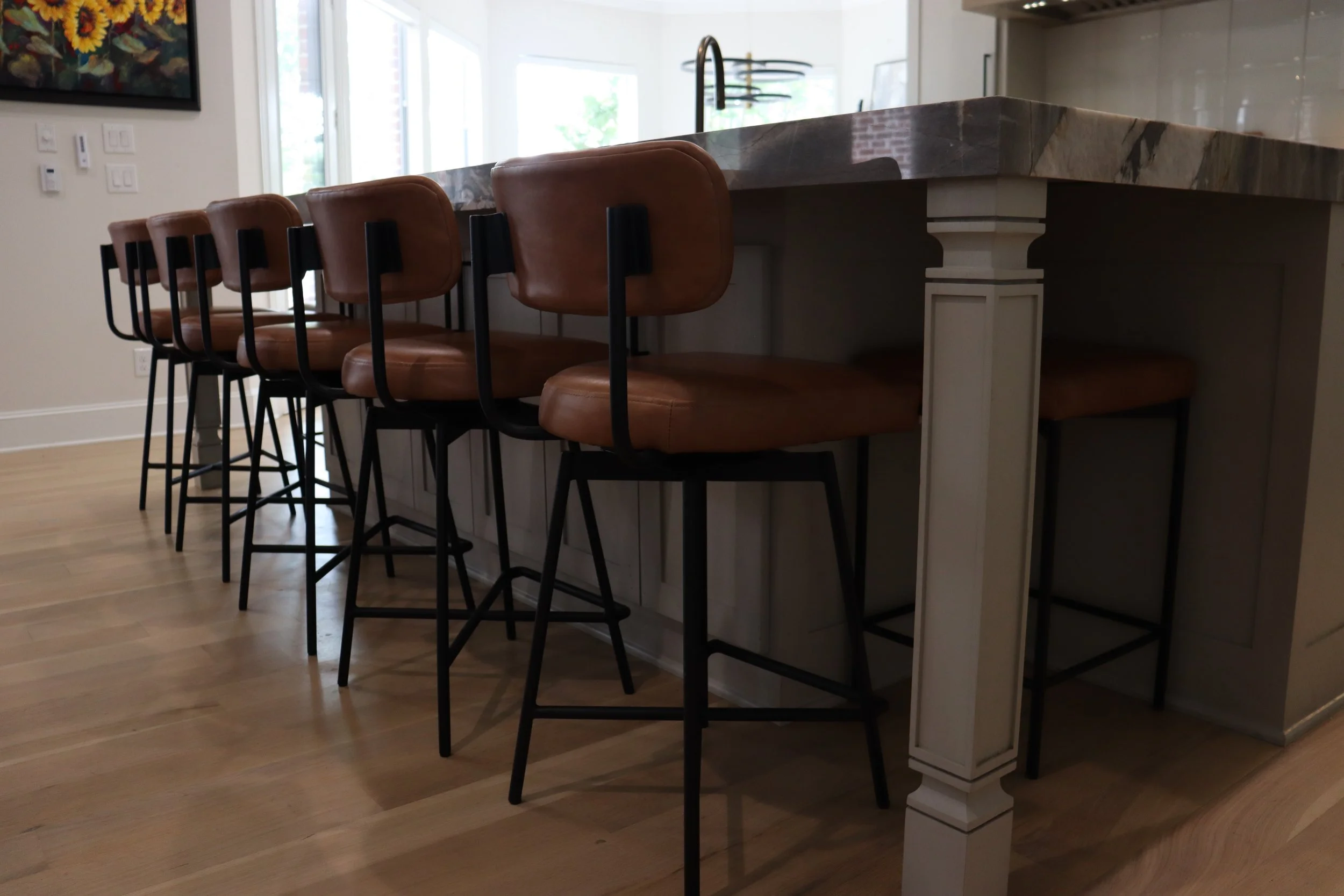 Interior view of a kitchen island with brown leather barstools and a marble countertop in a well-lit space with large windows.