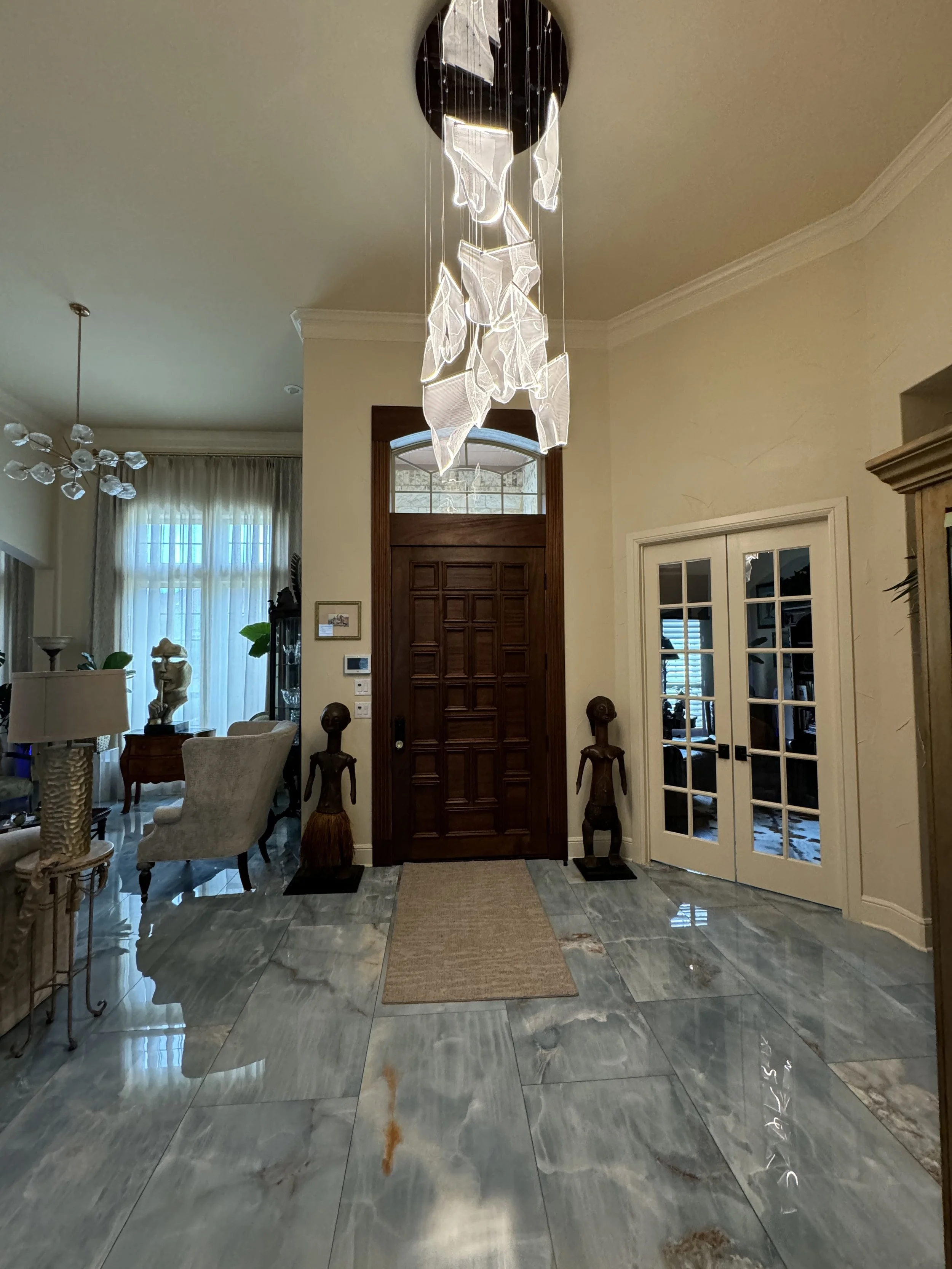 A modern front hall with a decorative chandelier hanging from the ceiling and a large wooden front door. The floor is made of polished marble tiles, and there is a beige rug in the center. On the left side, there are some chairs and sculptures, while on the right side, there are glass-paneled double doors leading to another room.
