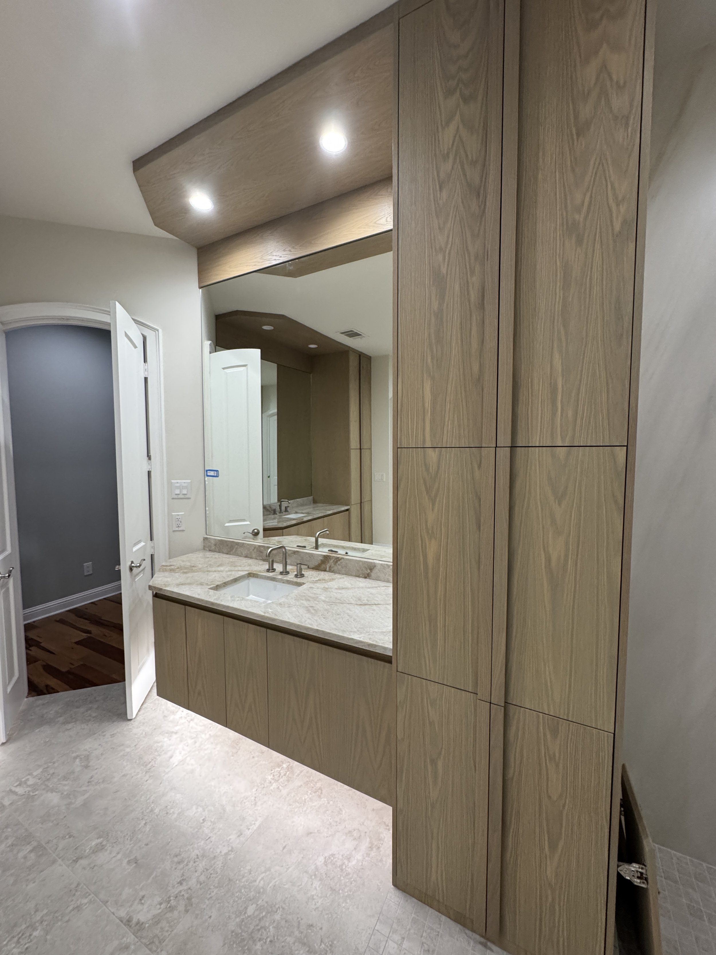 Interior view of a bathroom with a large mirror, wooden cabinets, and a marble countertop with two sinks. The bathroom has a sloped ceiling with recessed lighting and an open door leading to another room.